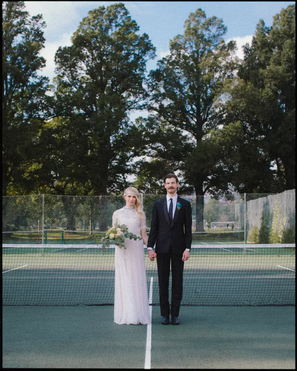 Bride in a beaded blush dress and groom in a dark suit hold hands, standing centred on a green tennis court