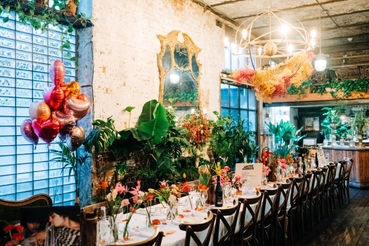 A long, elegant dining table is set with flowers and glassware in a rustic, plant-filled room at Clapton Country Club. Heart-shaped balloons and a chandelier create a festive, cozy atmosphere perfect for an East London wedding.