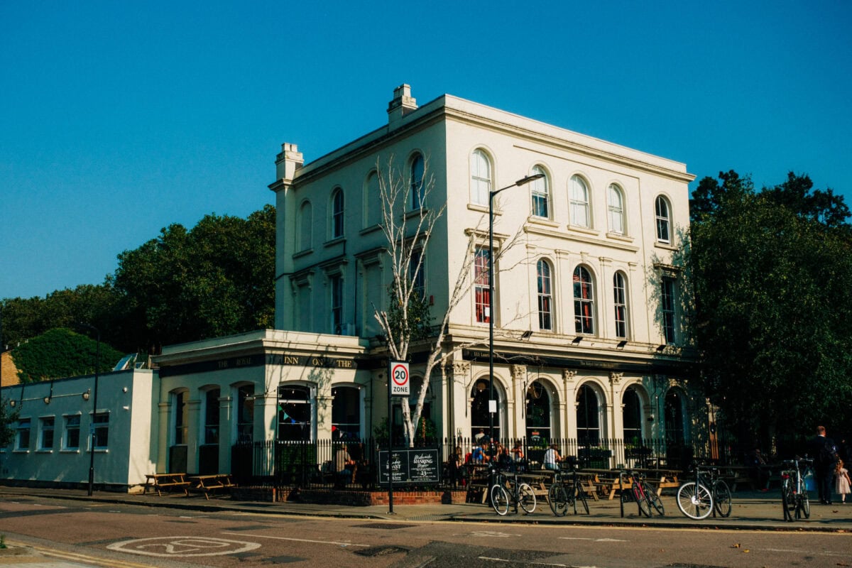 A large, white, three-story building with arched windows houses a lively outdoor pub the Royal inn on the pakr near Hackney Town Hall. People sit at tables under clear blue skies, bicycles line the fence, and trees surround this inviting East London spot.