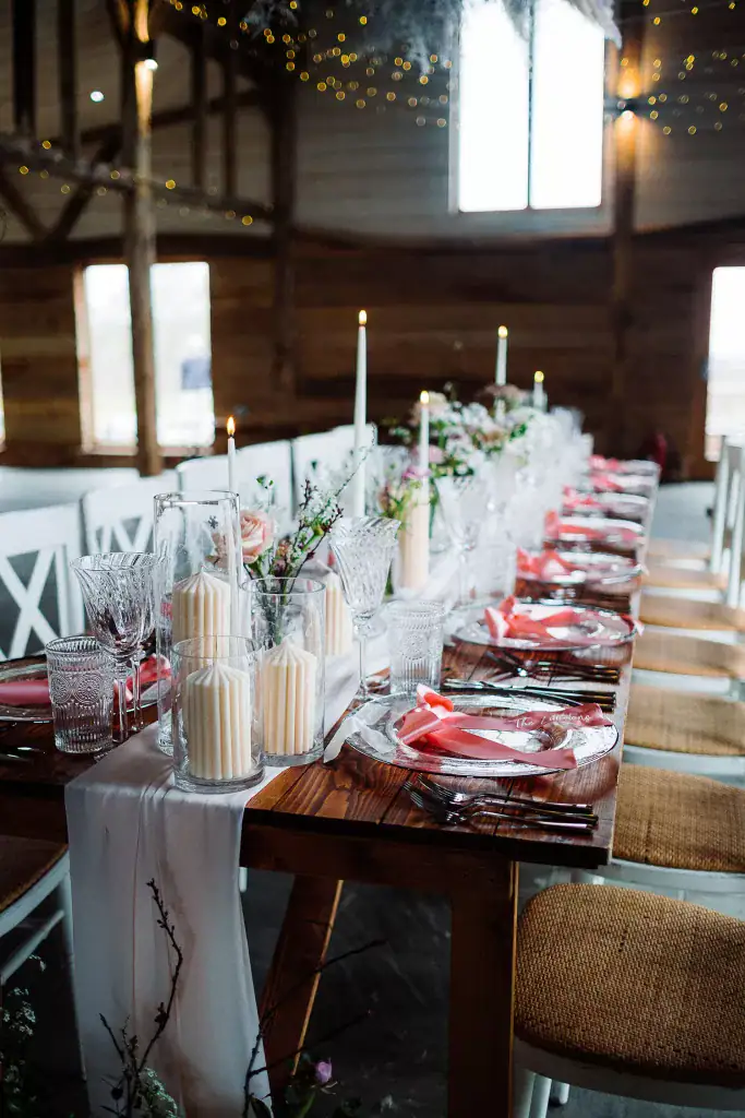 A rustic wooden table set for a formal event with candles, glassware, pink napkins, white chairs, floral arrangements, and soft fairy lights overhead—capturing the charm of a rustic barn wedding in a cozy cherry barn rye setting.