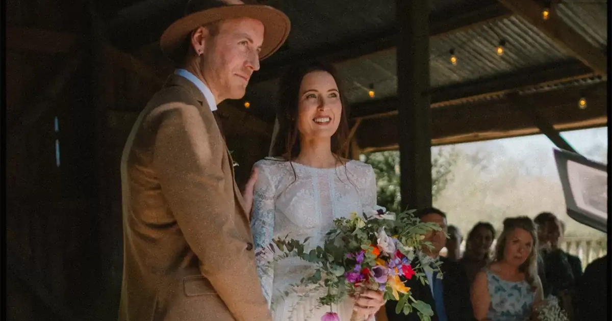 A bride in a white dress holding a colorful bouquet stands beside a groom in a brown suit and hat during a rustic indoor wedding ceremony—perfect inspiration for those who love rustic barn weddings—with guests seated in the background.