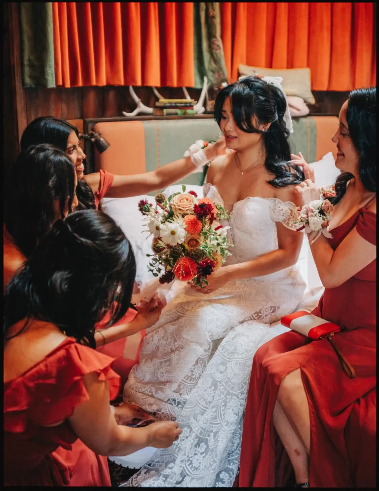 A bride in a white lace gown holds a bouquet while sitting on a bed at the autumn wedding bell in Ticehurst, surrounded by four bridesmaids in matching rust-colored dresses, all smiling and chatting in a warmly lit room.