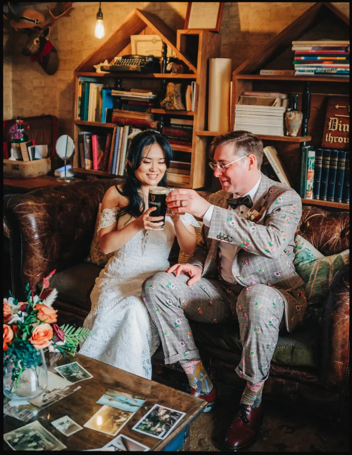 A bride in a white dress and groom in a patterned suit sit on a couch at The Bell in Ticehurst, smiling and clinking glasses. Surrounded by books, flowers, and scattered photos, they enjoy an autumn cosy wedding in a warmly lit room.