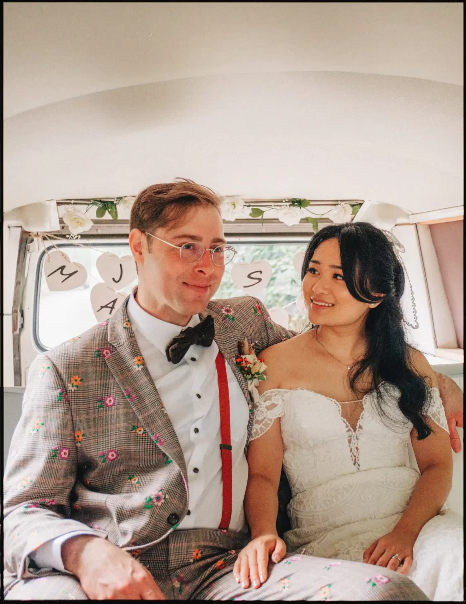 A bride in a white lace dress and a groom in a floral-patterned suit sit together in a decorated vehicle at their Autumn Wedding Bell in Ticehurst, smiling at each other with heart-shaped signs hanging behind them.