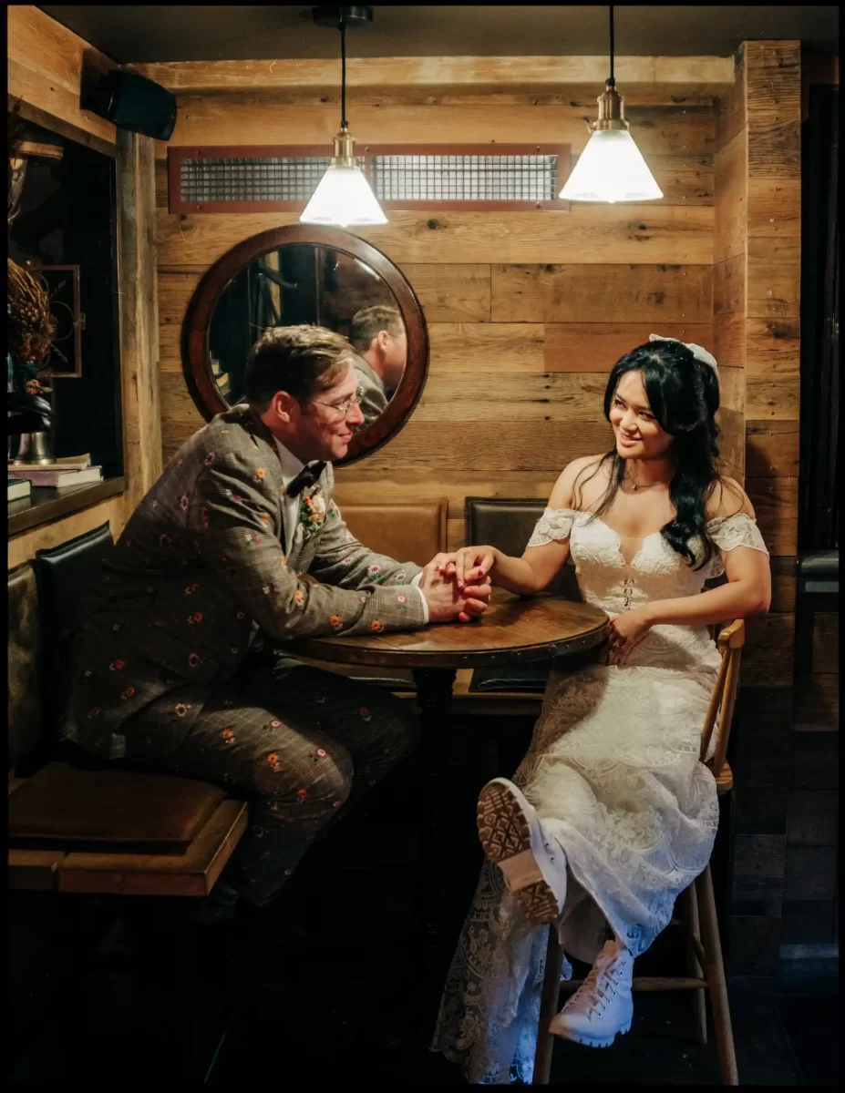A bride in a white dress and boots sits at a small wooden table holding hands with her groom in a patterned suit. They share a warm smile in the cozy, rustic glow of an autumn wedding at Bell in Ticehurst.