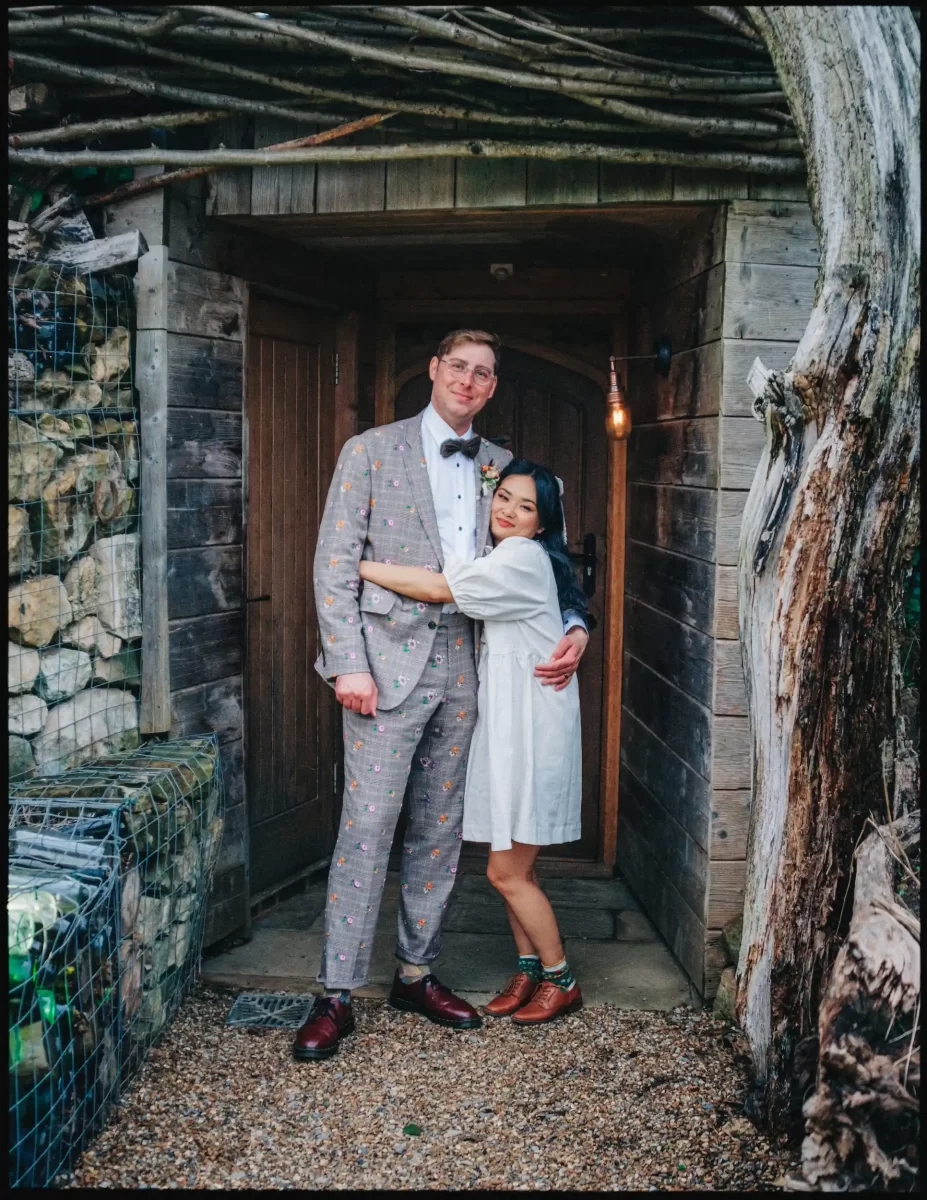 A tall man in a patterned suit and bow tie stands smiling with a shorter woman in a white dress and colorful shoes, hugging him in front of a rustic wooden doorway at Autumn Wedding Bell in Ticehurst, with stone walls and branches overhead.