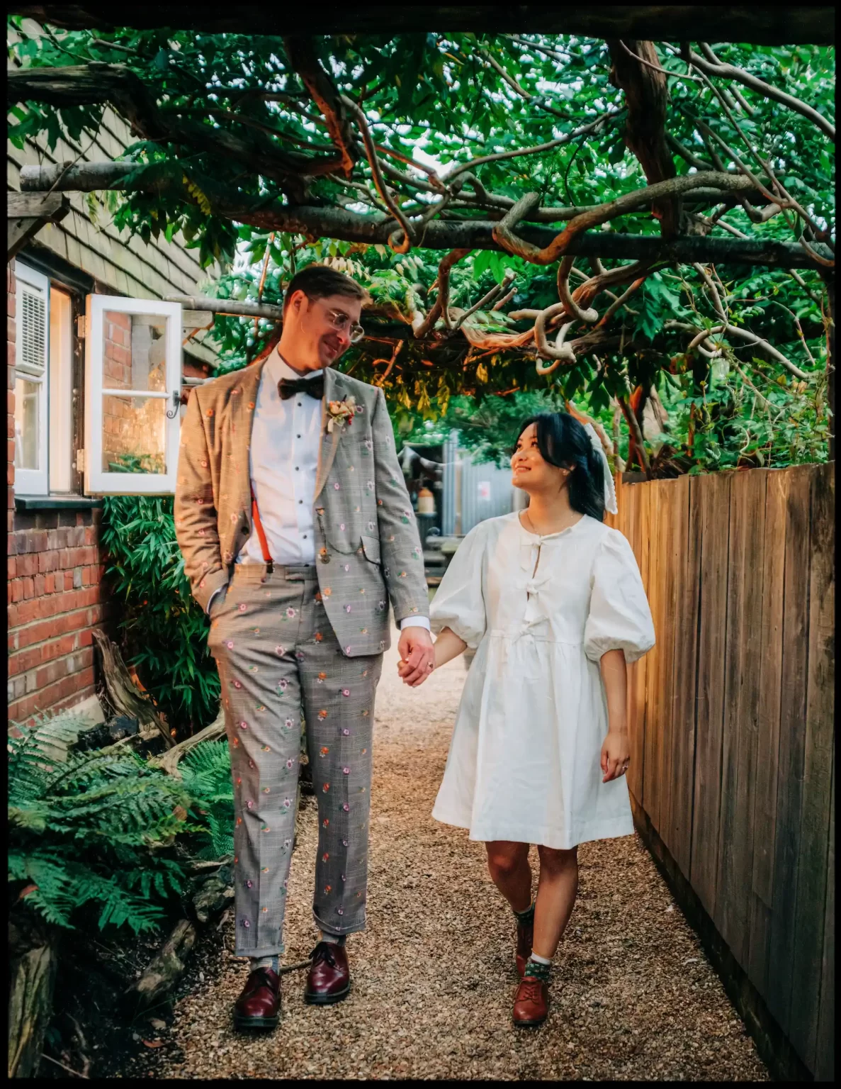 A couple holding hands and smiling at each other while walking outdoors under green foliage at an autumn wedding bell in Ticehurst. The man wears a patterned gray suit and bow tie, while the woman wears a white dress with puffed sleeves.