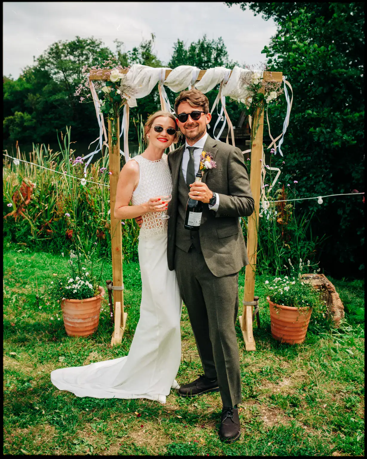 A bride in a white dress and a groom in a suit stand smiling under a rustic outdoor arch at Frickley Lake, Ninfield East Sussex, holding drinks, surrounded by greenery and potted plants.
