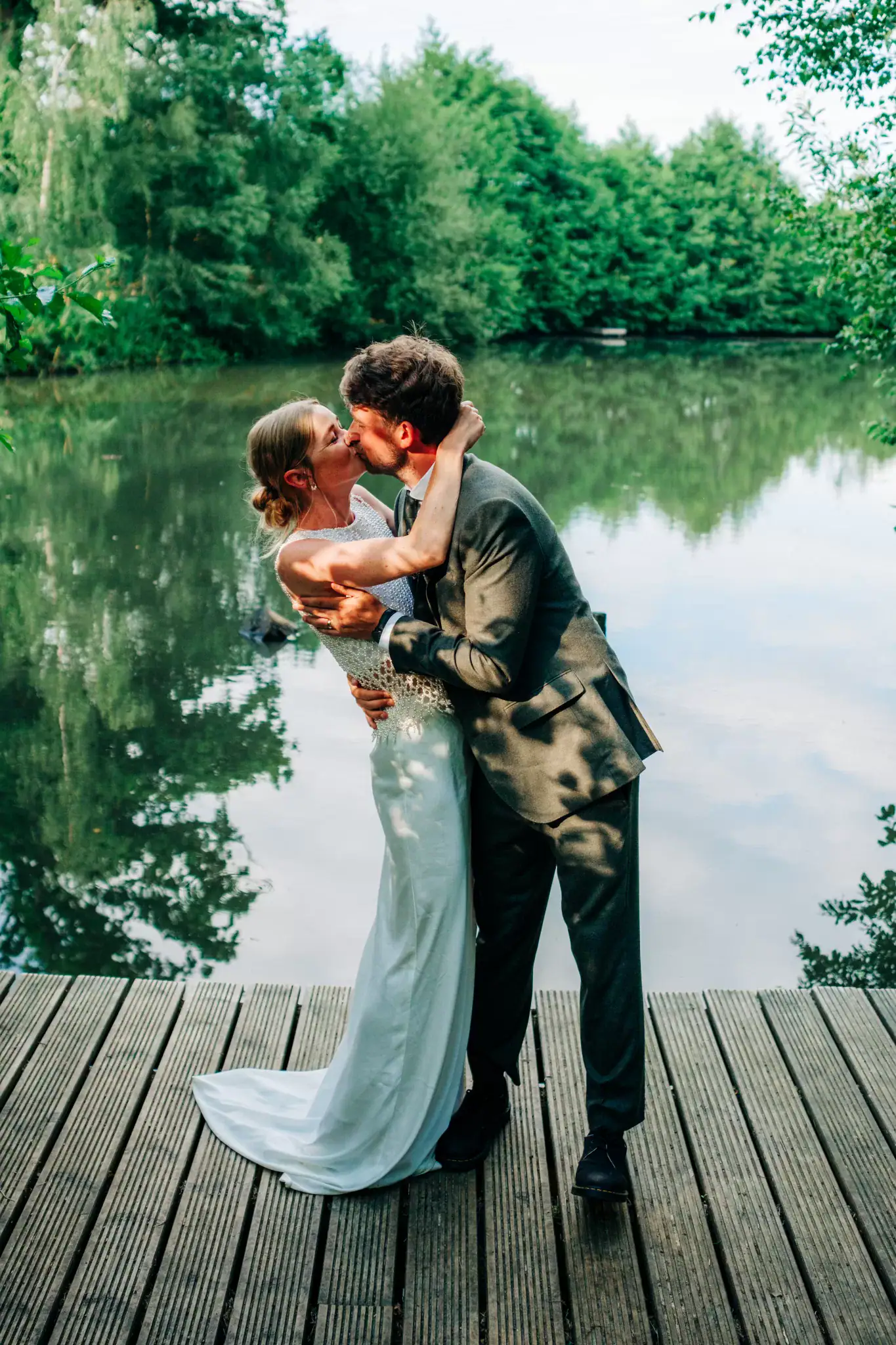 A couple dressed in wedding attire kisses on a wooden dock beside a calm lake at Frickley Lake wedding venue near Battle, Sussex, surrounded by lush green trees and reflected in the water on a sunny, festival-like day.