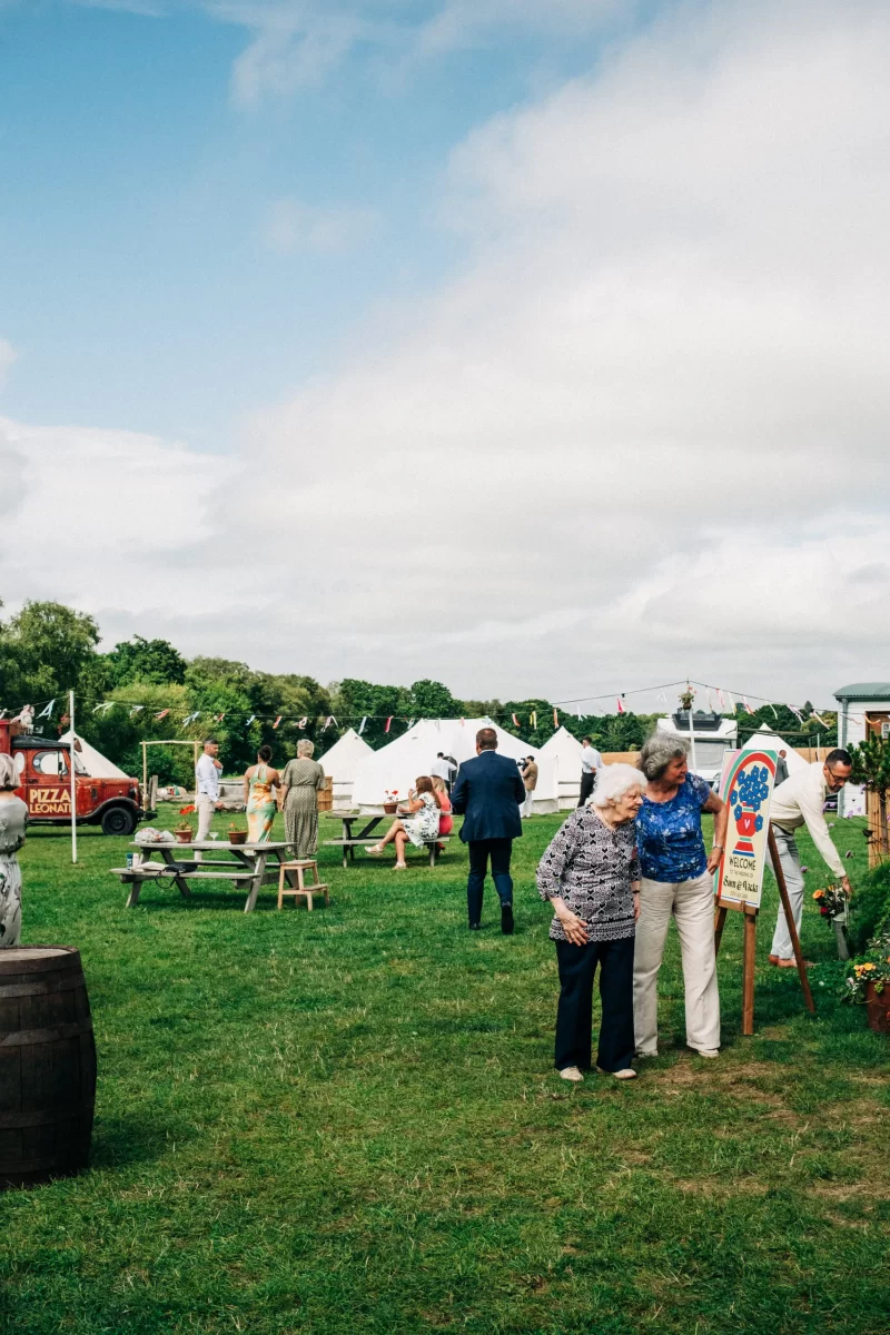Two older women look at a sign at an outdoor fair. People sit at picnic tables near white tents on a grassy field under a partly cloudy sky, creating a setting full of relaxed festival wedding vibes and garden festival wedding ideas.