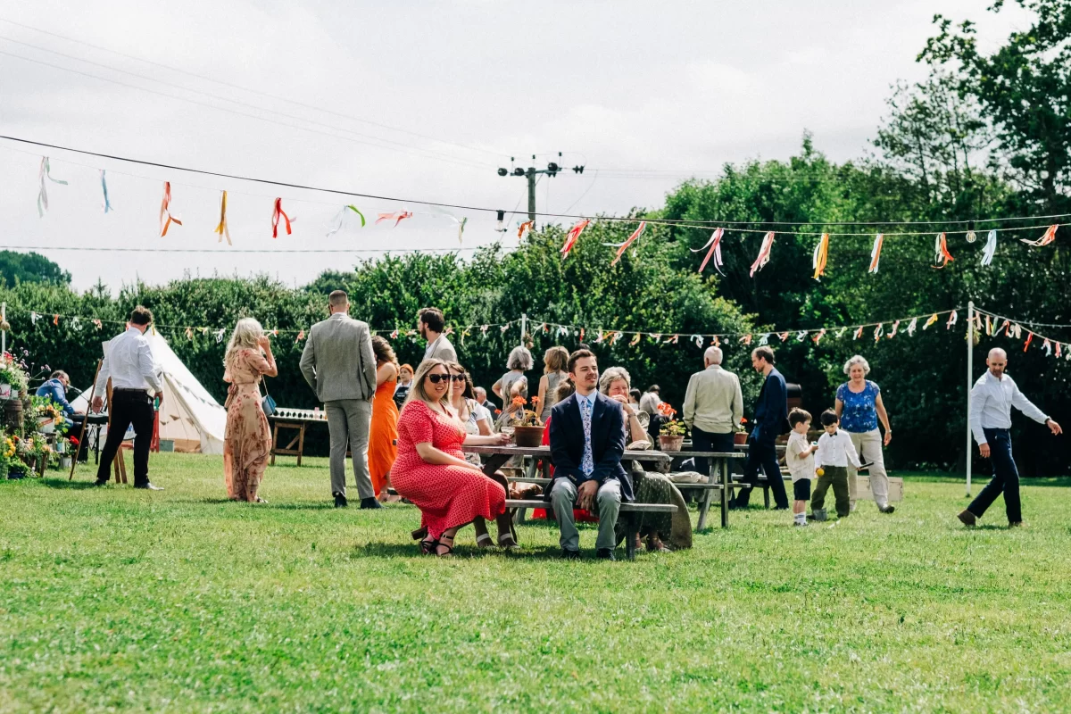 A group of people socialize at a Relaxed Festival wedding on a sunny day. Colorful ribbons are strung overhead, with some guests sitting at wooden tables and others strolling the grass—perfect inspiration for garden festival wedding ideas.