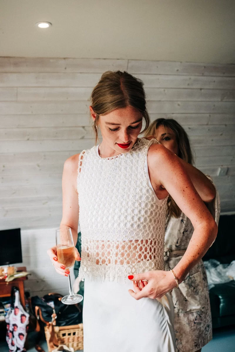 A woman in a sleeveless white textured top and skirt holds a champagne glass and looks down, adjusting her outfit—capturing the relaxed festival wedding vibe. Another woman stands behind her in a softly lit room with light wood walls.