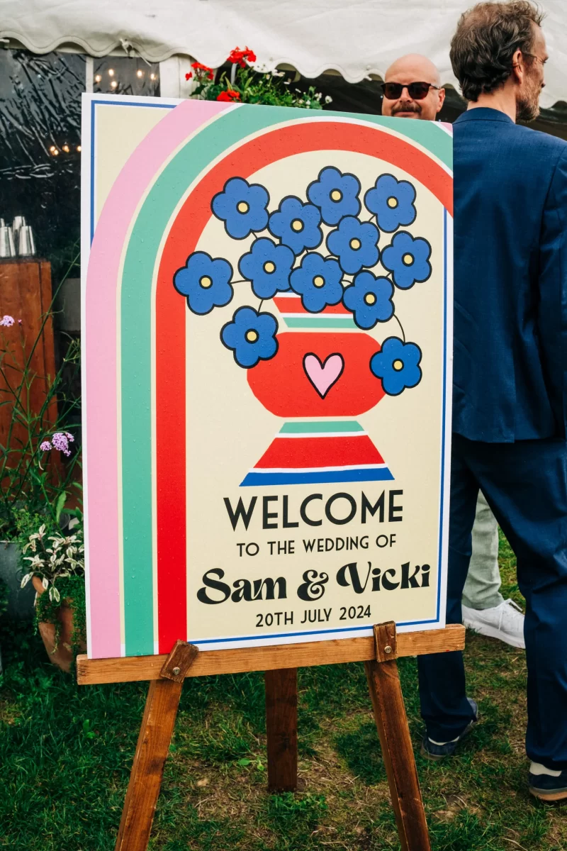 A colorful wedding welcome sign captures relaxed festival wedding vibes with blue flowers in a red vase, a pink heart, rainbow arches, and text: Welcome to the wedding of Sam & Vicki, 20th July 2024. People and garden festival wedding ideas appear in the background.
