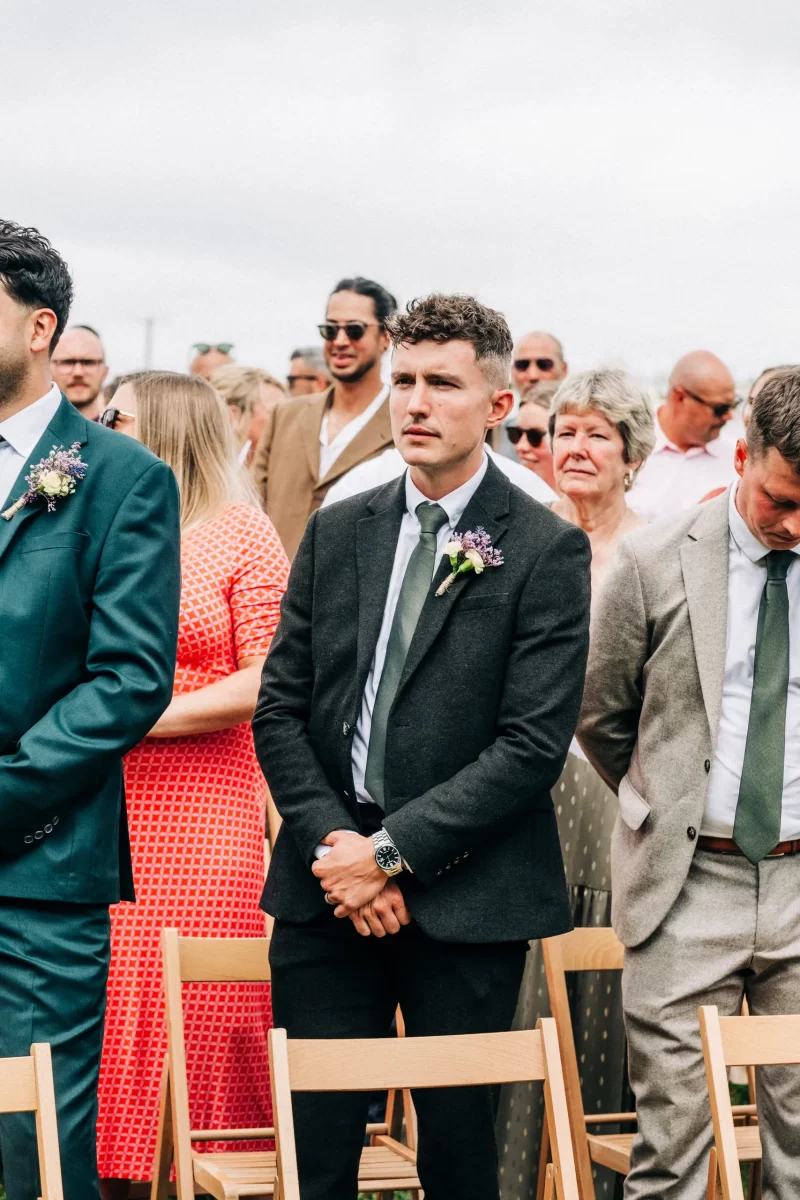 A man in a black suit with a boutonniere stands among other guests at a relaxed festival wedding. Surrounded by people in formal attire and sunglasses, he enjoys the garden festival wedding atmosphere and the charm of wooden chairs outdoors.