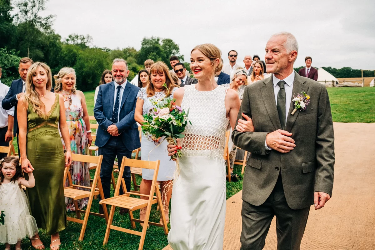 A bride in a white dress and a man in a suit walk arm in arm outdoors, smiling, while guests stand and watch. Relaxed Festival wedding vibes fill the air with empty wooden chairs, green grass, and trees in the background.