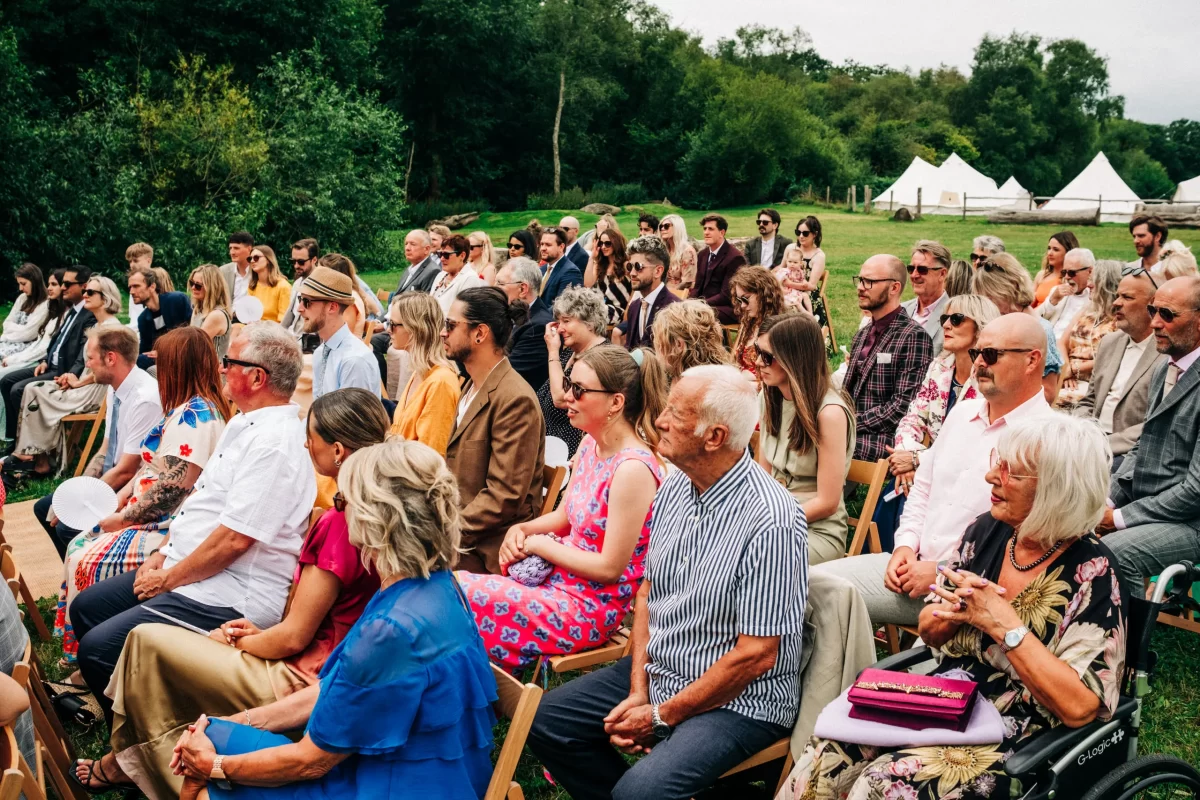 A large group of people sits outdoors on wooden chairs, dressed in summer clothing, attentively watching a relaxed festival wedding. Green grass, trees, and white tents add inspiration for garden festival wedding ideas in the background at Frickley Lake near Battle