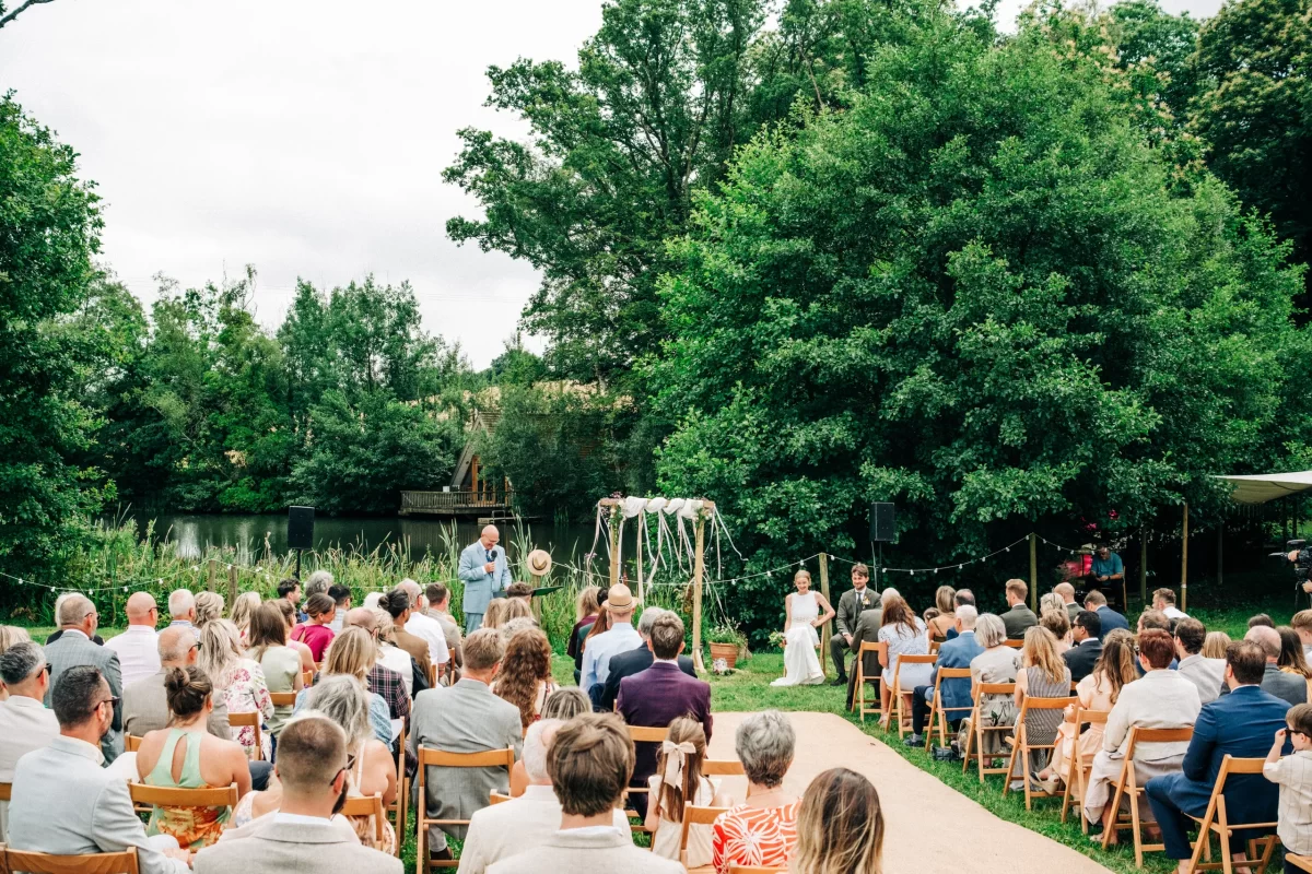 Outdoor wedding ceremony by a lake, with guests seated on wooden chairs facing a couple and officiant under a floral arch, surrounded by green trees—perfect for relaxed festival wedding vibes and inspiring garden festival wedding ideas.