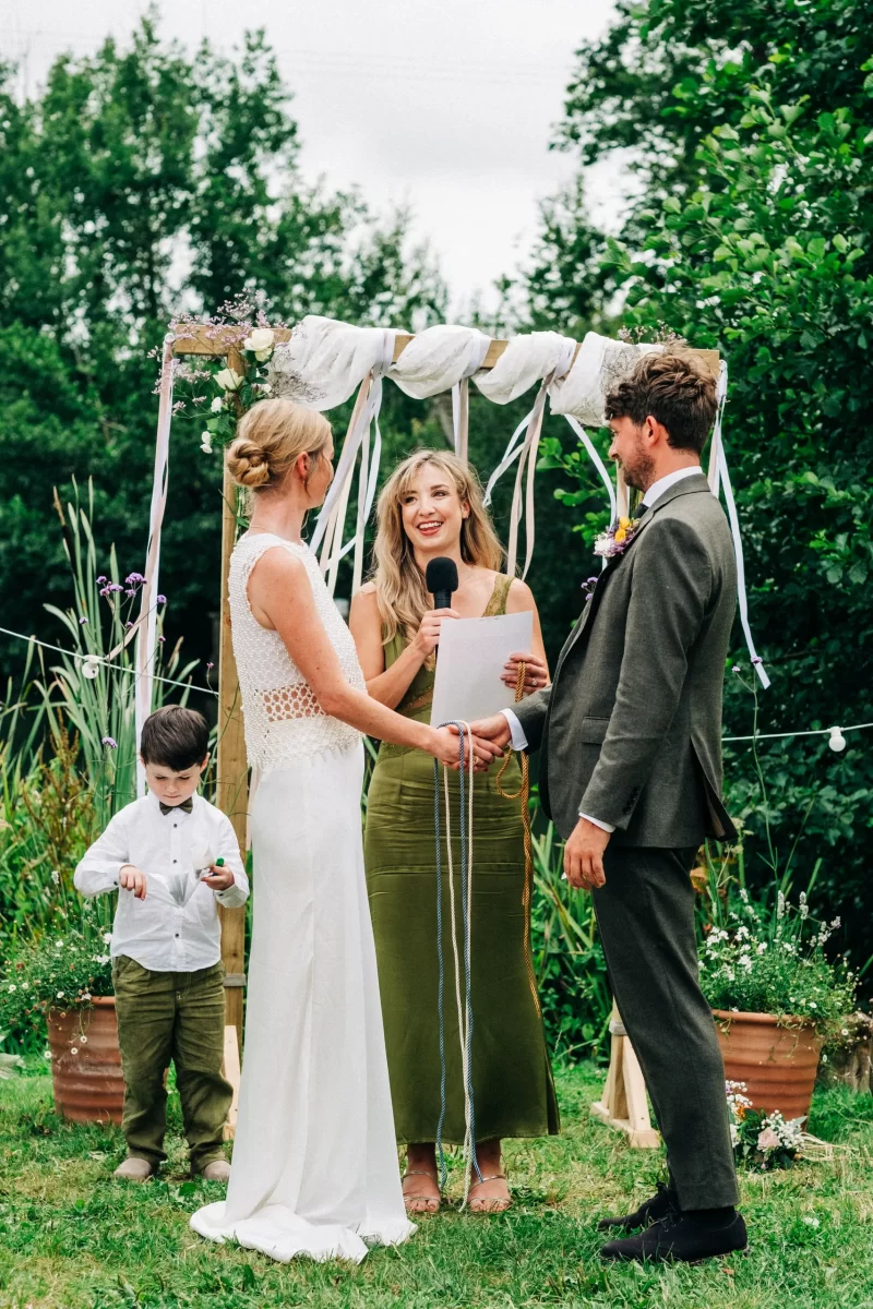 A bride and groom hold hands and face each other during a relaxed festival wedding ceremony. An officiant stands between them with a microphone and script, smiling, as a young boy looks down nearby. Green foliage surrounds this garden festival wedding scene.