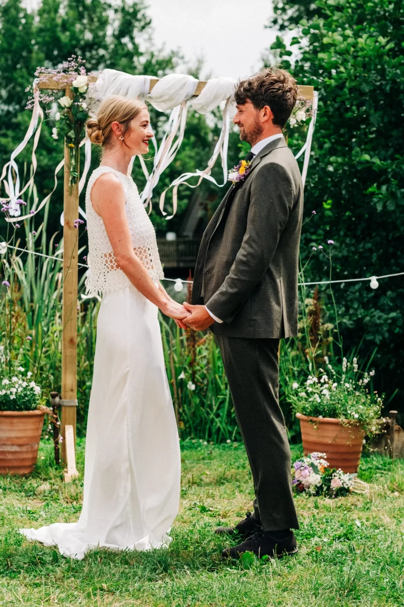 A bride and groom stand facing each other, holding hands and smiling outdoors. Embracing relaxed festival wedding vibes, they are surrounded by greenery, potted flowers, and a rustic wooden arch adorned with white ribbons.