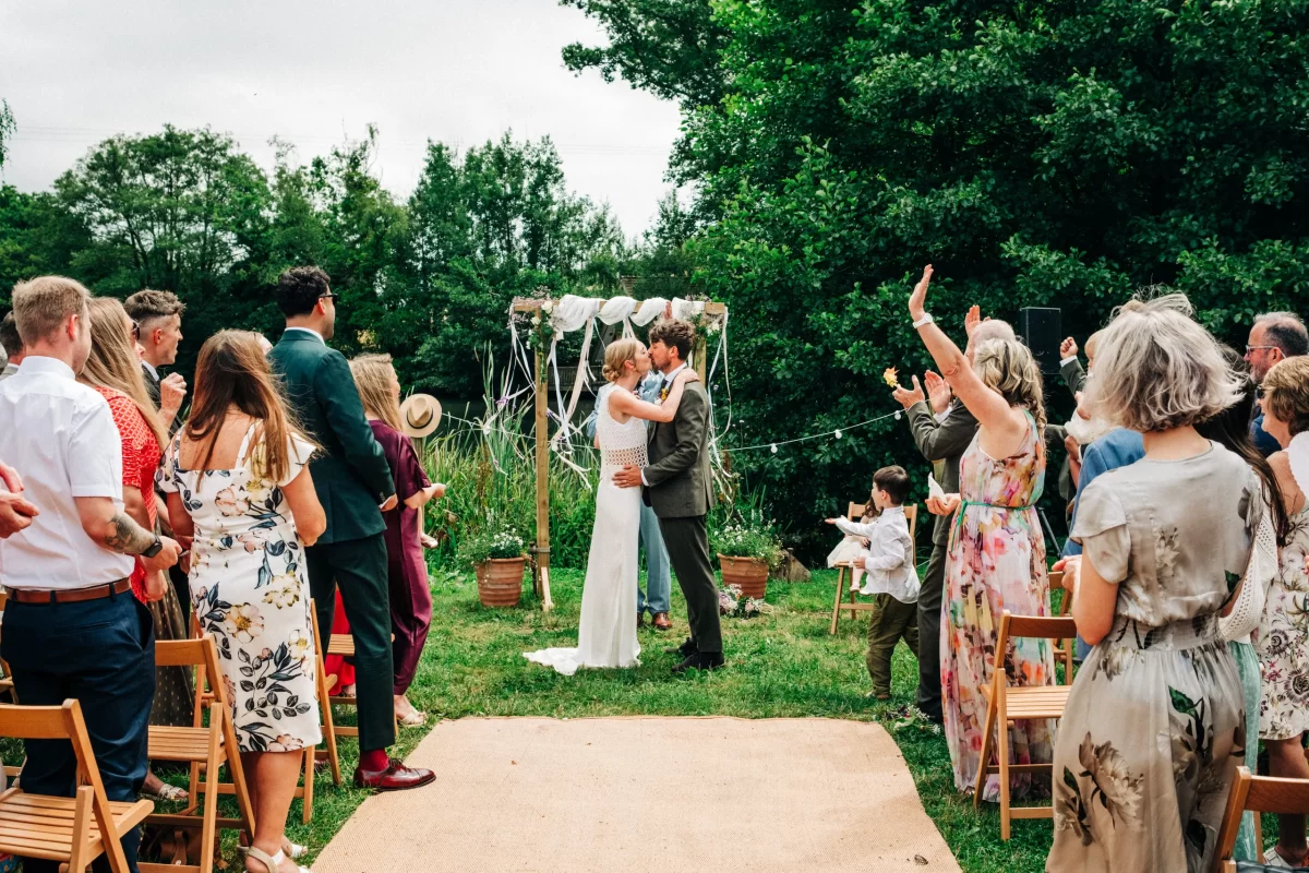 A bride and groom kiss at a relaxed festival wedding ceremony as guests in summer attire applaud. The lush green setting, with trees and a decorated arch, inspires beautiful garden festival wedding ideas.