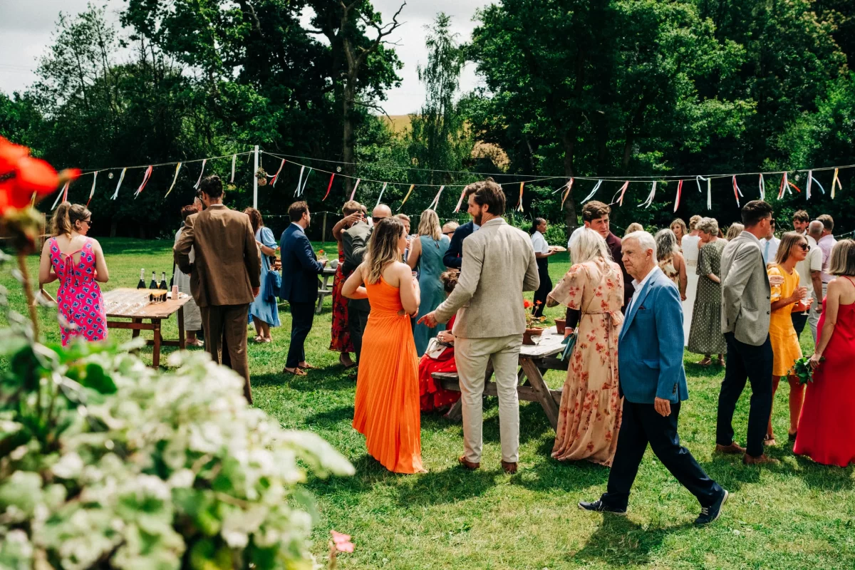A group of people in colorful formal attire gather outdoors on grass, with tables, bunting, and trees in the background, capturing the essence of a Relaxed Festival wedding and offering inspiration for garden festival wedding ideas.