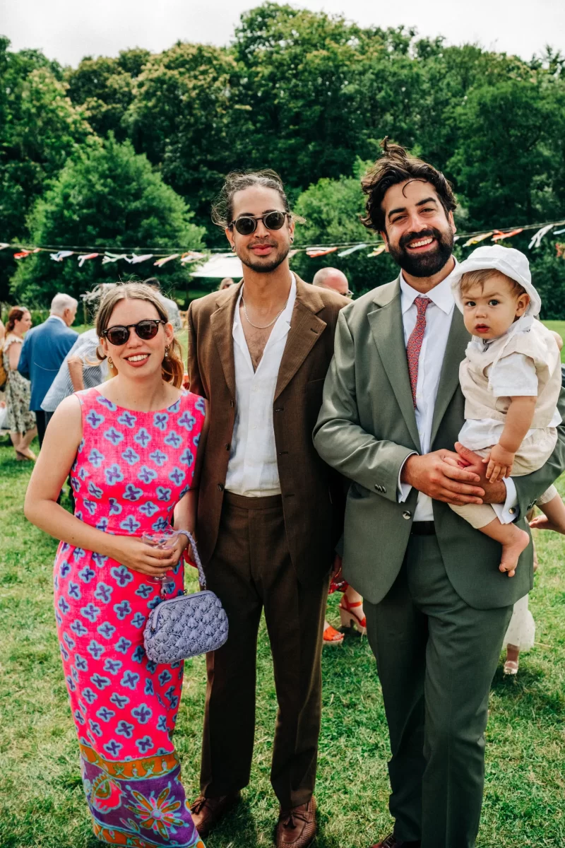 Three adults and a baby stand on grass at an outdoor event, evoking relaxed festival wedding vibes. The woman wears a bright pink patterned dress, while the men in brown and green suits pose with trees in the background—perfect garden festival wedding ideas.