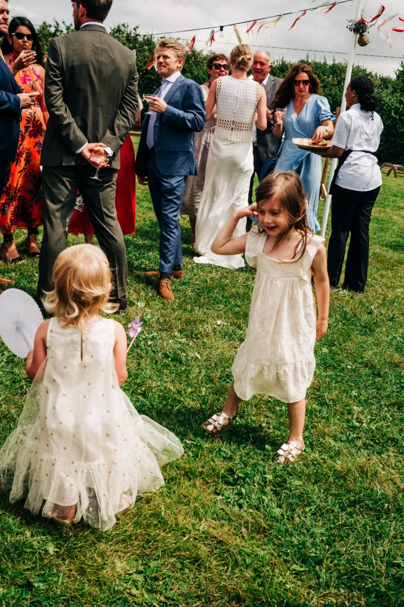 Two young girls in white dresses play on grass at a Relaxed Festival wedding, while adults in formal attire chat under a decorated canopy. The lively, summery scene captures charming garden festival wedding ideas.