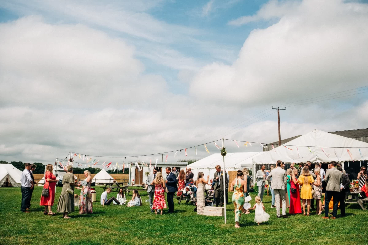 A group of people in colorful clothing gather outdoors on a grassy field for a relaxed festival wedding celebration, with white tents, picnic tables, and bunting flags—perfect inspiration for garden festival wedding ideas under a partly cloudy sky.