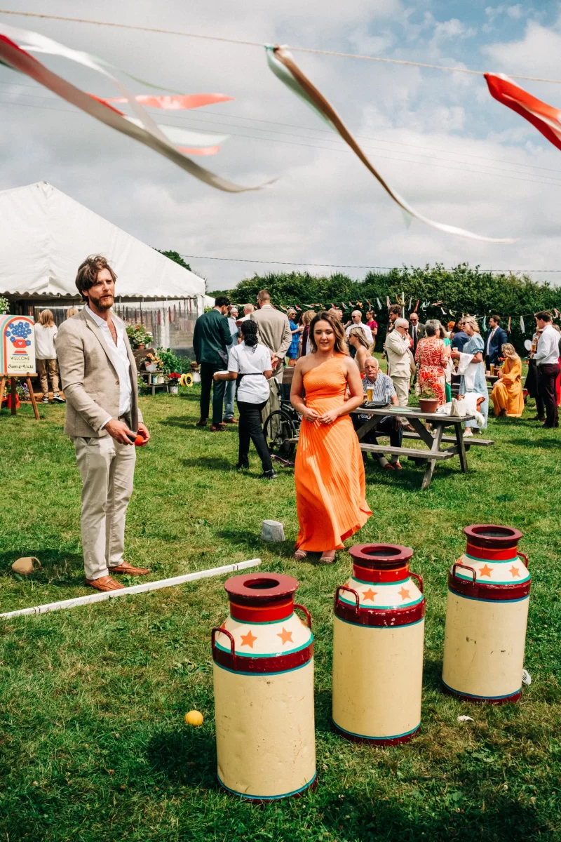 A man in a tan suit and a woman in an orange dress play a carnival game with large decorated milk cans at a relaxed festival wedding, while a crowd gathers near a white tent. Colorful ribbons hang above, adding to the garden festival wedding ideas.