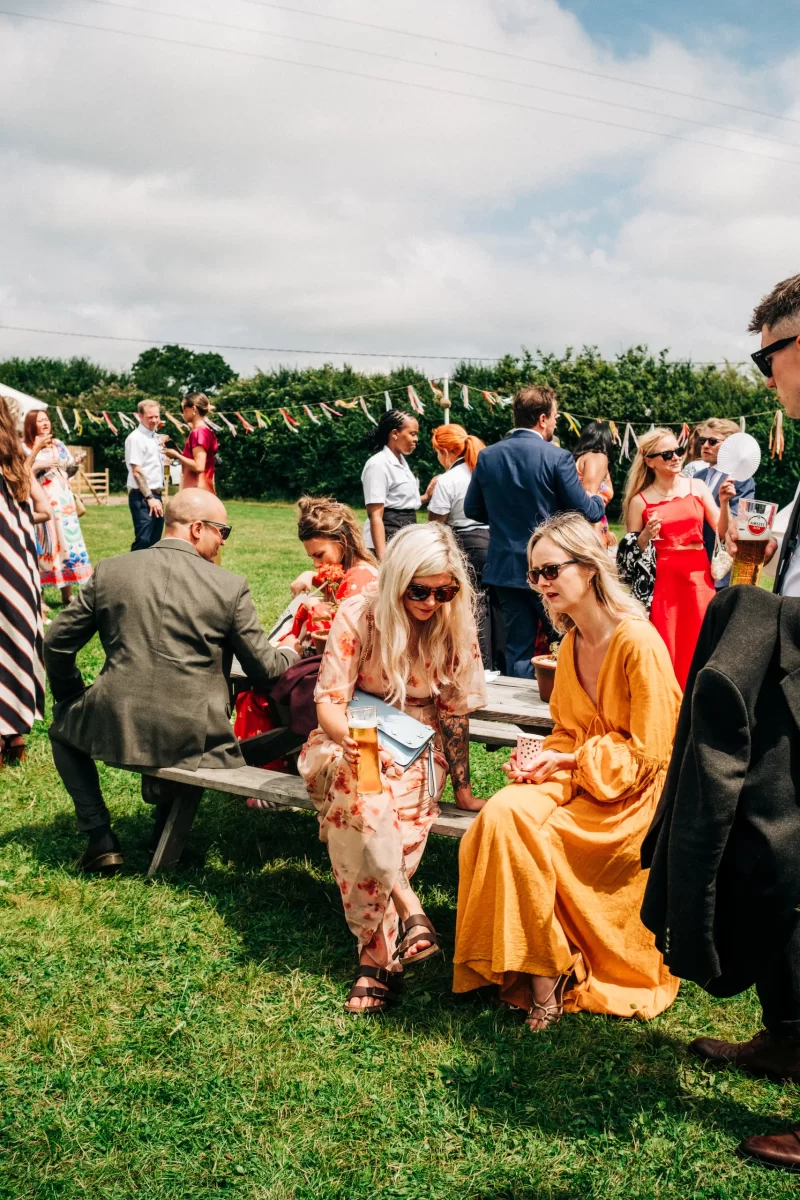 A group of people at a relaxed festival wedding, some sitting on picnic benches and others standing on the grass. Two women in colorful dresses chat in the foreground under sunny skies, with garden festival wedding ideas visible in the cheerful decorations.