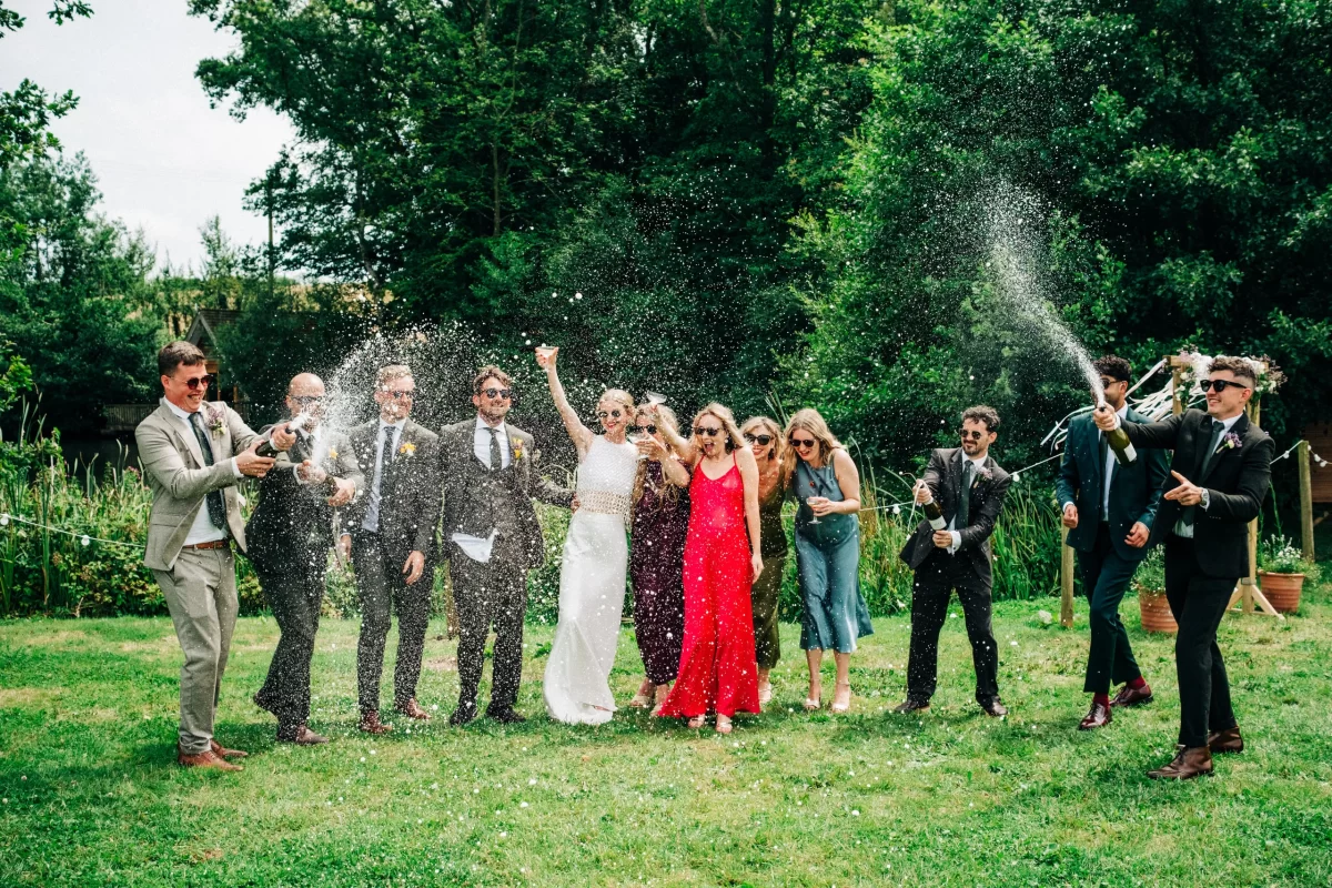 A group of people dressed formally are celebrating outdoors, spraying champagne and smiling joyfully at a relaxed festival wedding, with trees and garden festival wedding ideas in the background.