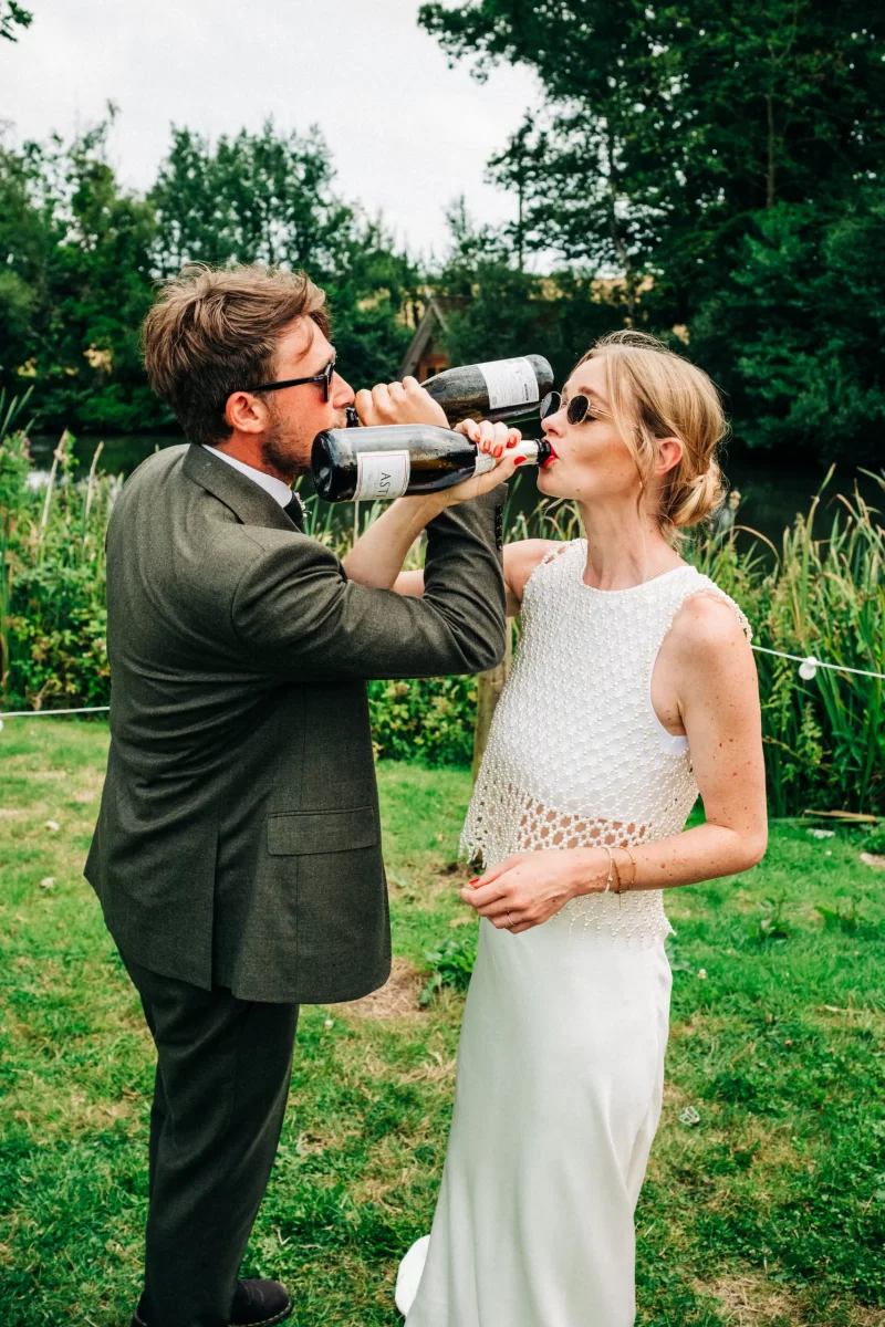 A man in a suit and a woman in a white dress and sunglasses drink from wine bottles together outdoors, surrounded by greenery—perfect inspiration for relaxed festival wedding vibes or unique garden festival wedding ideas.