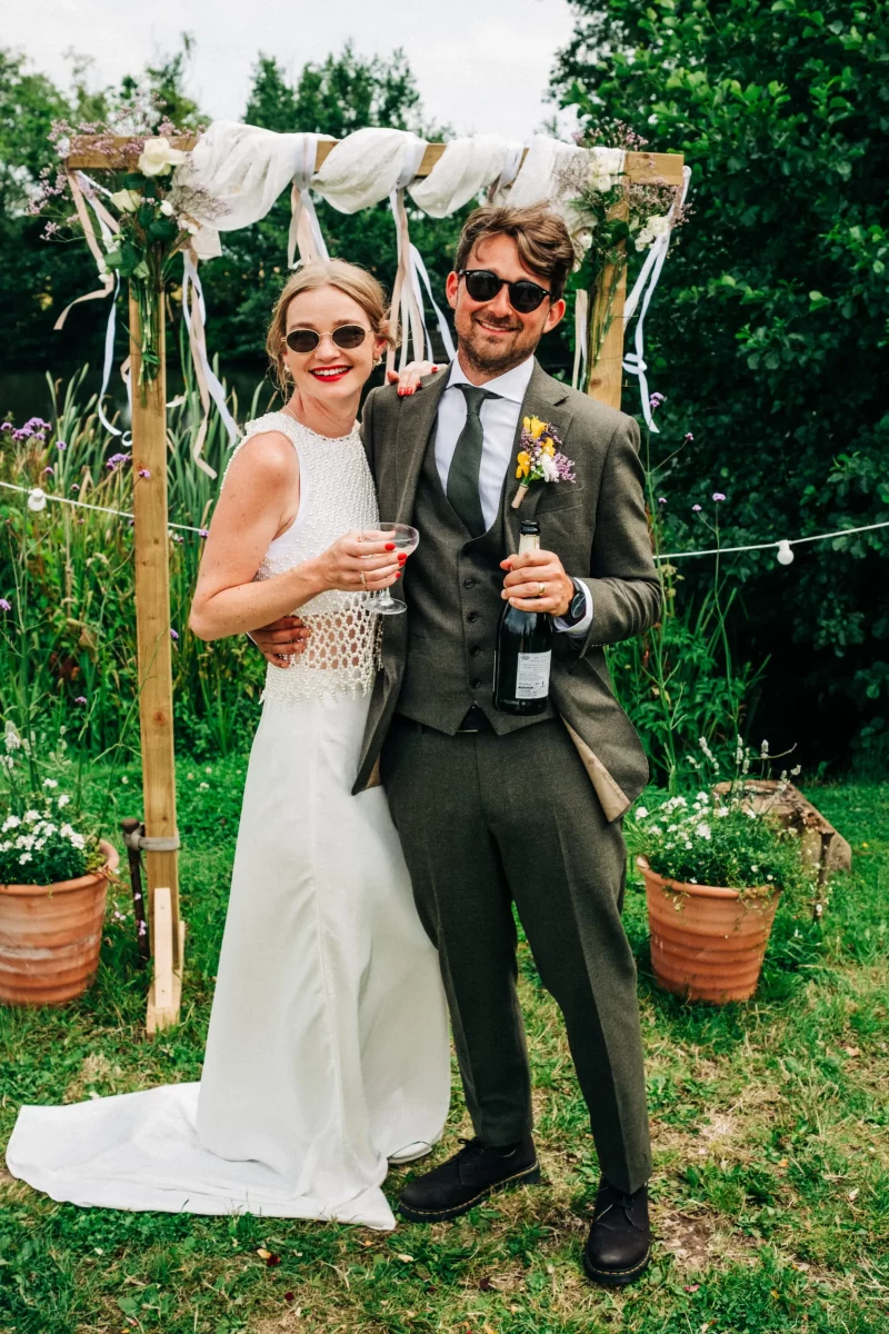 A smiling bride in a white dress and a groom in a green suit stand arm in arm outdoors, holding drinks, in front of a decorated wooden arch with greenery and flowers—a perfect scene for relaxed festival wedding inspiration. Both wear sunglasses and look happy.