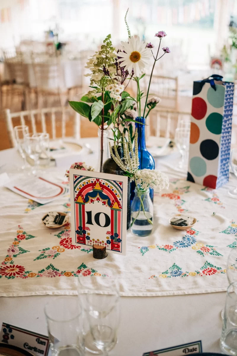 A decorated table set for a relaxed festival wedding features a floral tablecloth, glassware, blue vases with wildflowers, a colorful card displaying the number 10, and a polka dot gift bag in the background—perfect for garden festival wedding ideas.