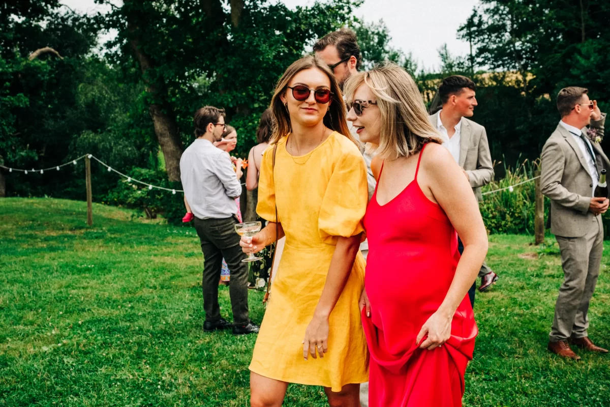Two women in bright dresses, one yellow and one red, walk together on a grassy lawn at a relaxed festival wedding. Other guests, including adults and a child, mingle in the background under trees on a cloudy day.