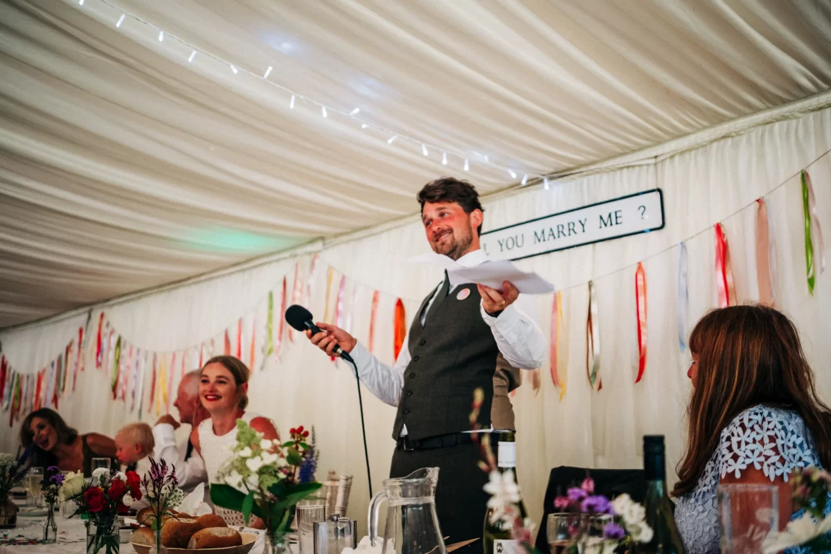 A man in a suit stands holding a microphone and paper, smiling and speaking at a decorated wedding reception table with flowers and guests, embodying Relaxed Festival wedding vibes under a sign reading YOU MARRY ME?.