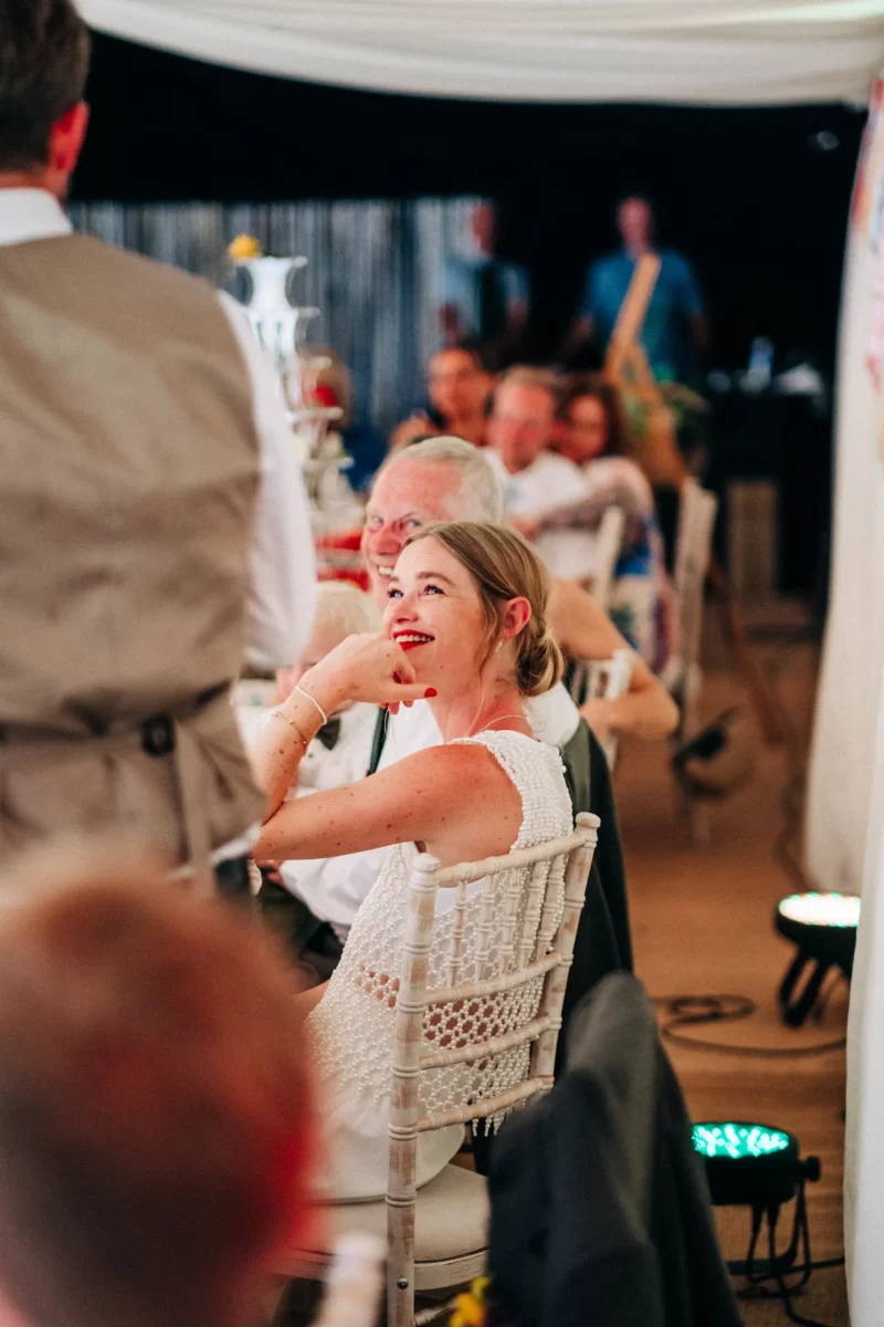 A woman in a white dress sits on a white chair at a decorated event, smiling and looking up at someone speaking, with other guests seated at tables in the background—perfect inspiration for relaxed festival wedding celebrations.