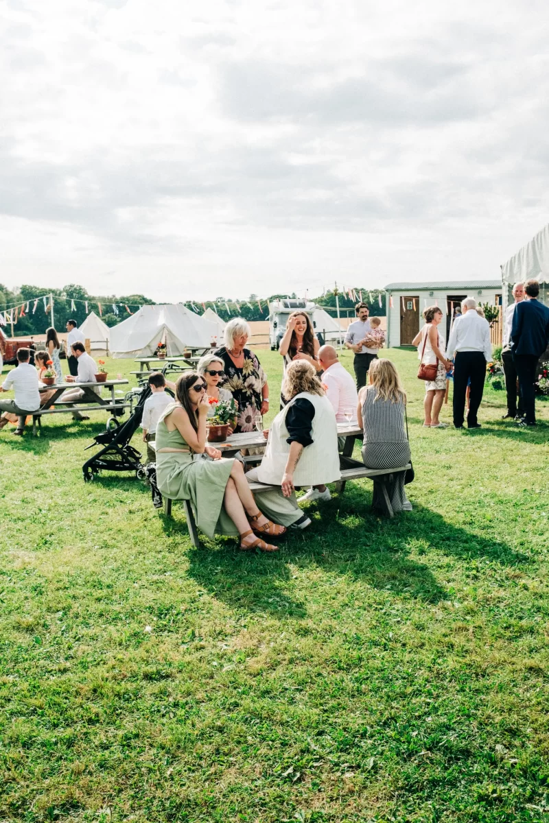 People relax and mingle around picnic tables at a garden festival wedding, with white tents and bunting in the background under a partly cloudy sky—perfect for gathering relaxed festival wedding ideas.