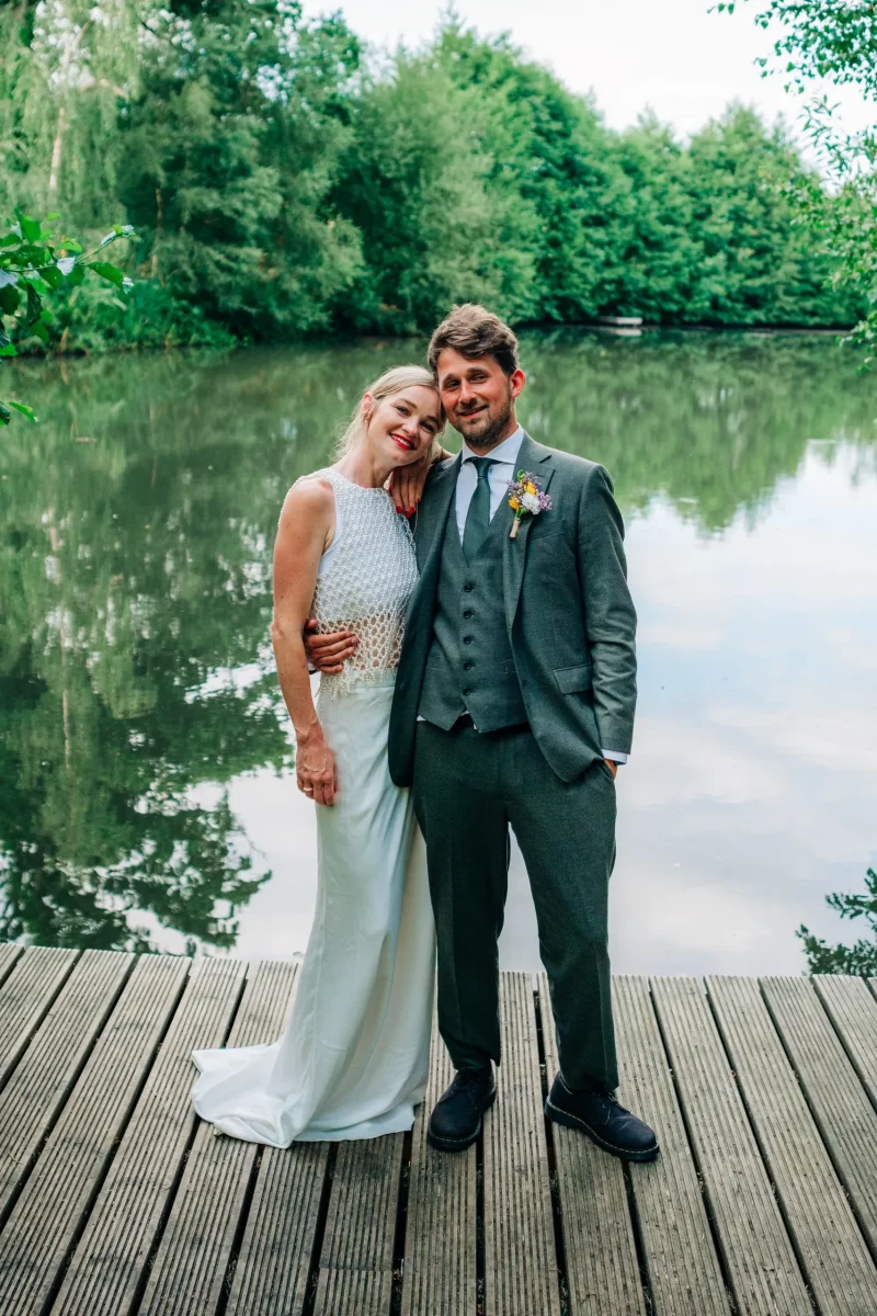 A bride in a white dress leans her head on the groom’s shoulder. The groom, in a gray suit, smiles as they stand on a wooden deck by a calm lake and green trees—a perfect scene for relaxed festival wedding inspiration.