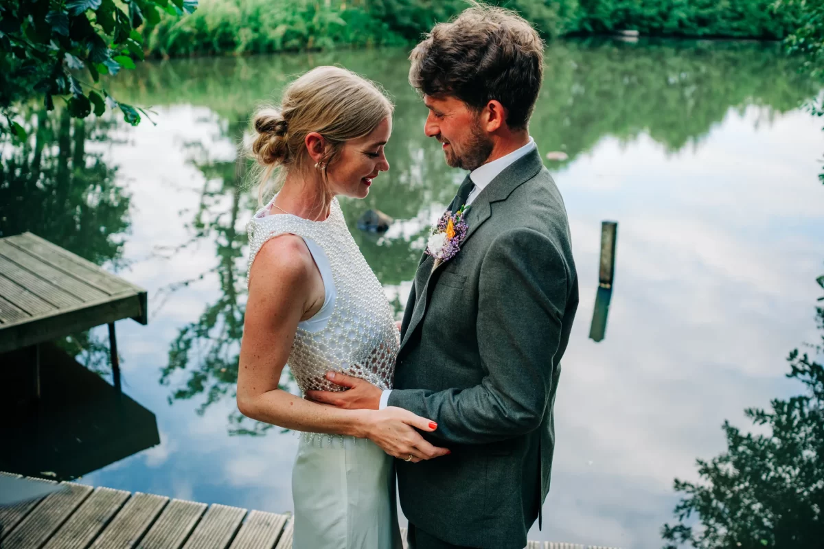 A bride and groom stand close, holding each other and smiling lovingly by a calm lakeside, surrounded by lush greenery—a perfect setting for relaxed festival wedding moments and inspiring garden festival wedding ideas.