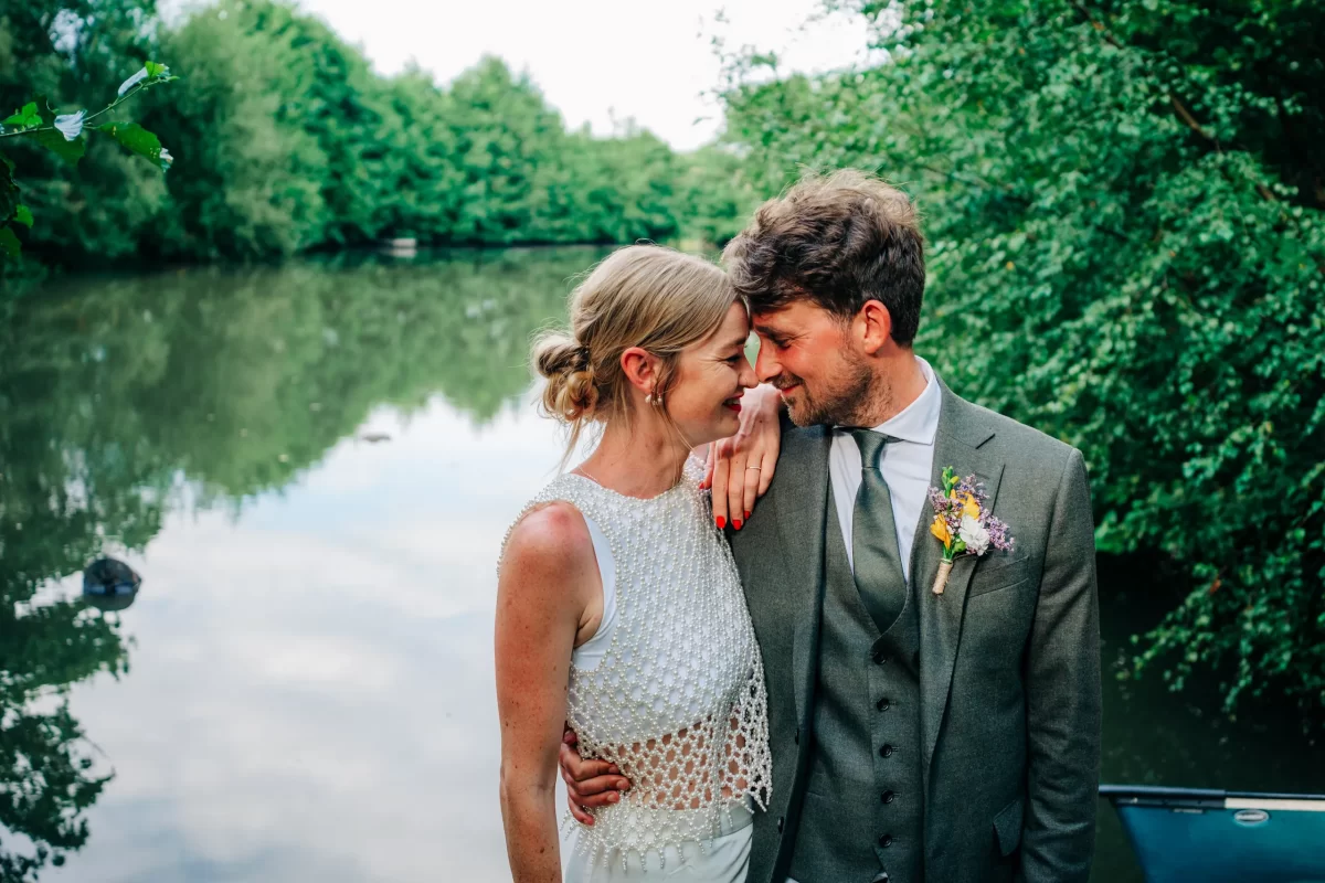 A smiling couple in wedding attire stand arm-in-arm by a calm lake, touching foreheads and surrounded by lush green trees—a perfect scene for relaxed festival wedding inspiration and garden festival wedding ideas.