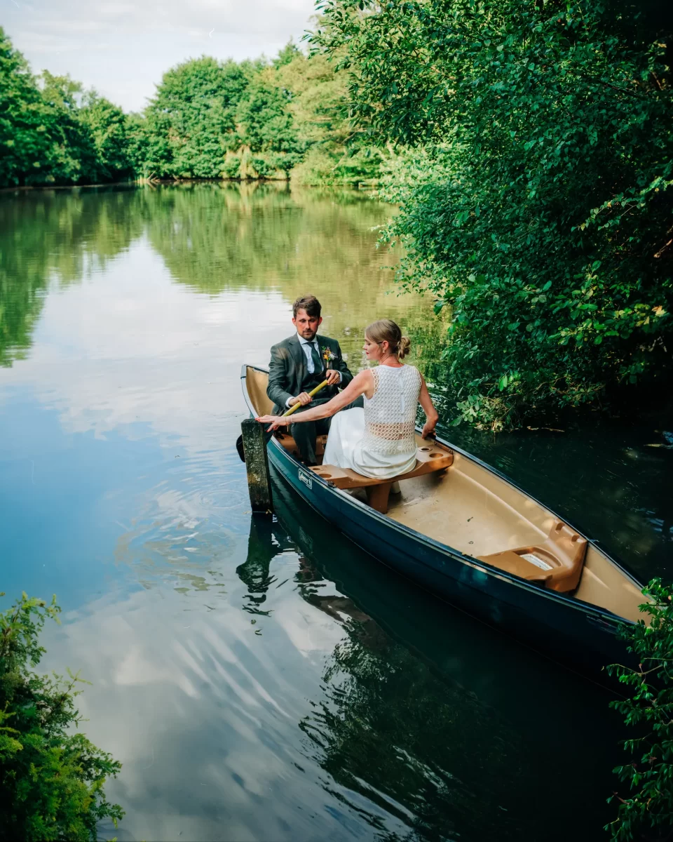 A man in a suit and a woman in a white dress sit facing each other in a canoe on a calm, green lake surrounded by trees—a serene scene perfect for garden festival wedding ideas under a bright sky.