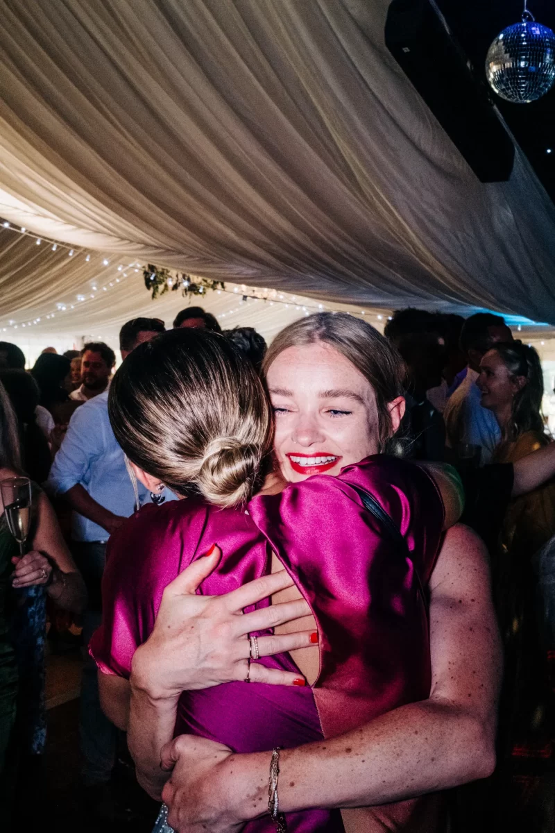Two women in vibrant magenta dresses embrace and smile joyfully at a lively, relaxed festival wedding, with string lights overhead. Other guests and a disco ball add to the festive indoor atmosphere.