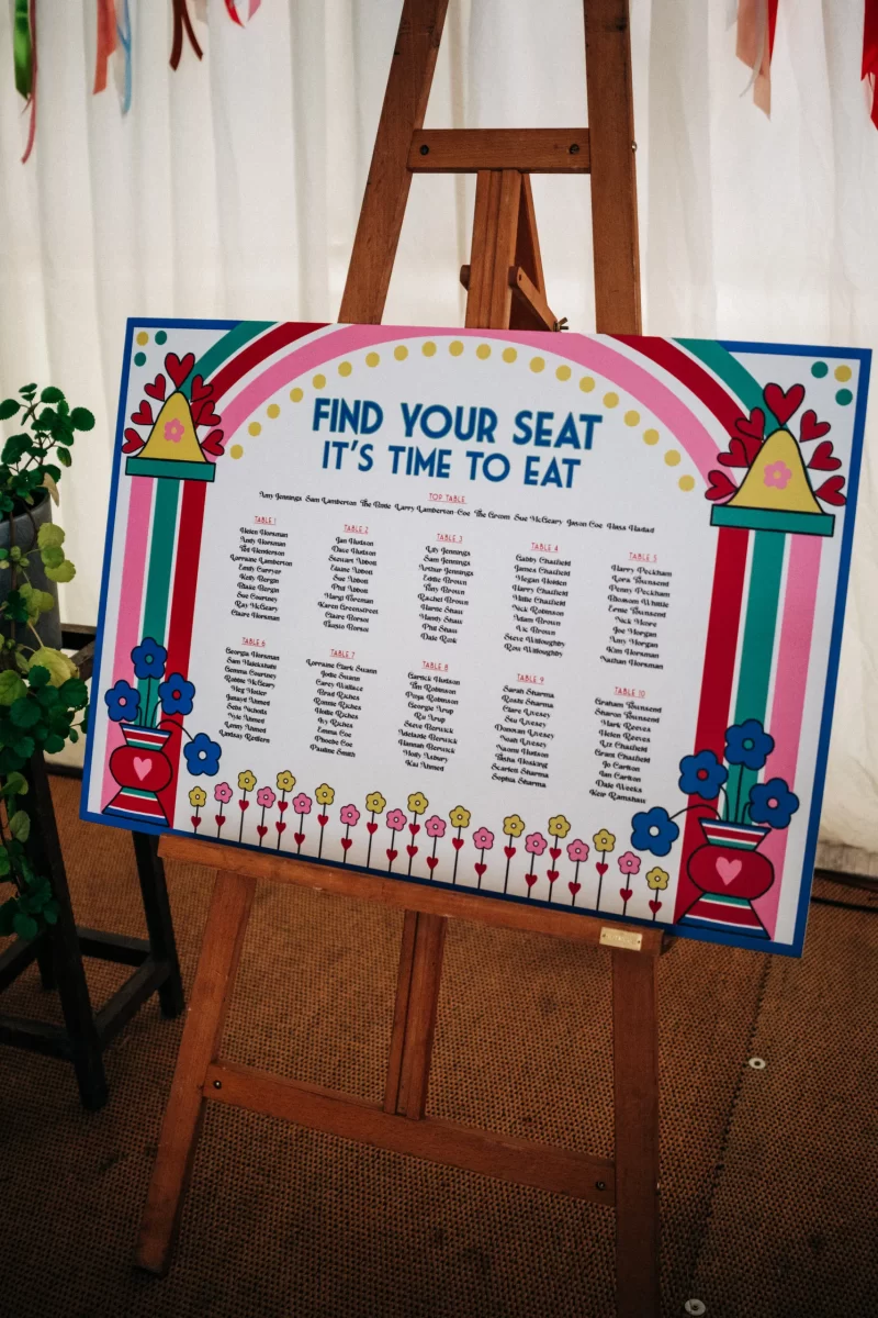 A colorful seating chart on an easel displays Find your seat, it's time to eat with tables and guest names, decorated with hearts, flowers, and rainbow patterns—perfect for relaxed festival wedding or garden festival wedding ideas.