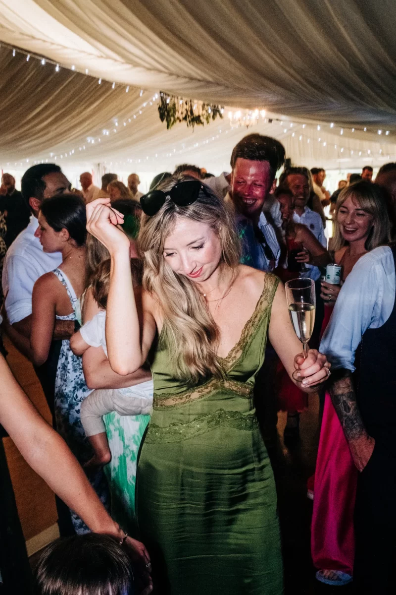 A woman in a green dress dances and smiles at a lively, crowded event under a tent, holding a glass of champagne—a perfect scene for those seeking garden festival wedding ideas or a relaxed festival wedding vibe.
