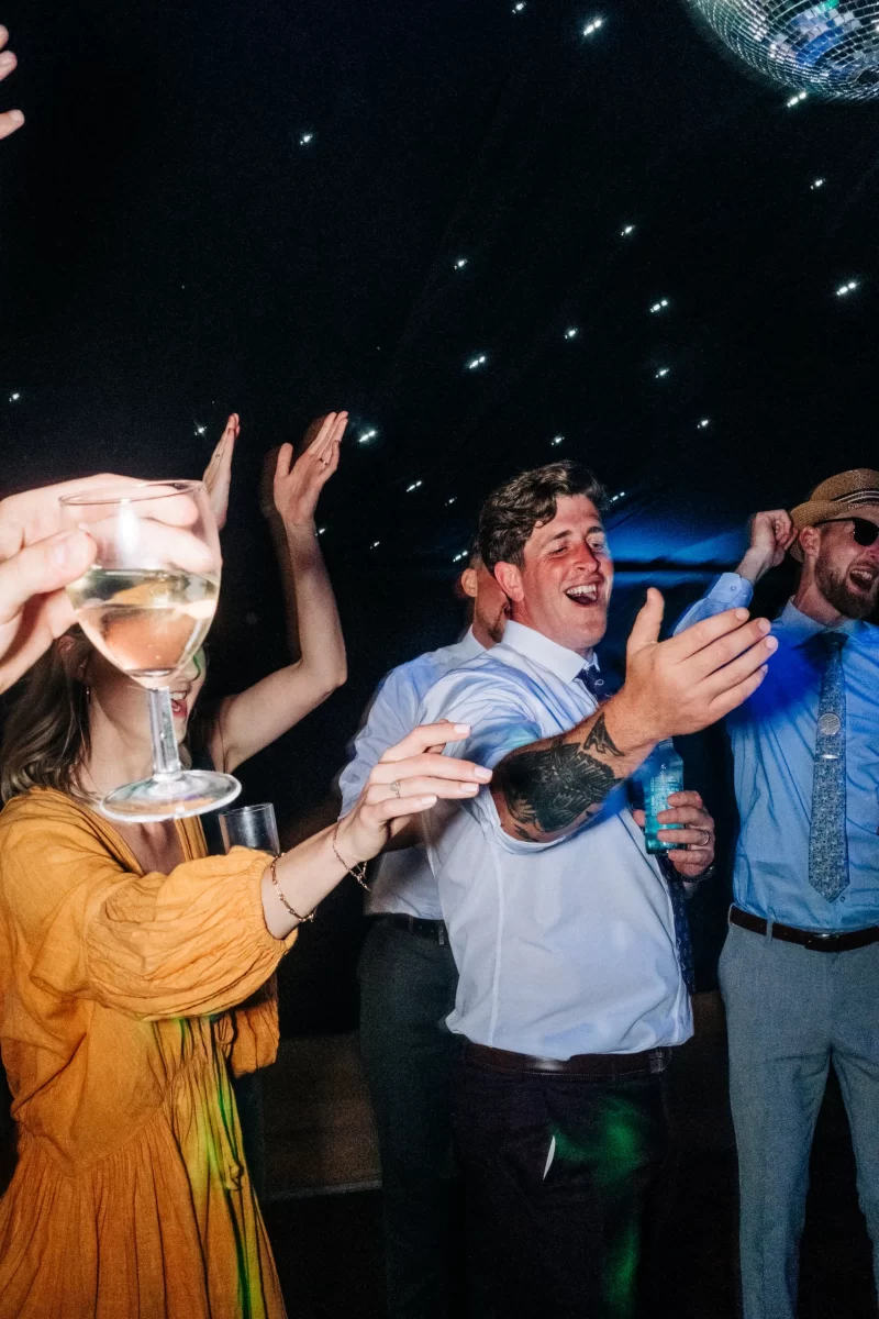 A group cheerfully dances at a Relaxed Festival wedding under star-like lights. A woman in a yellow dress holds white wine, while a man in a white shirt with tattooed arm leads the fun, smiling and reaching out. Perfect for garden festival wedding ideas.