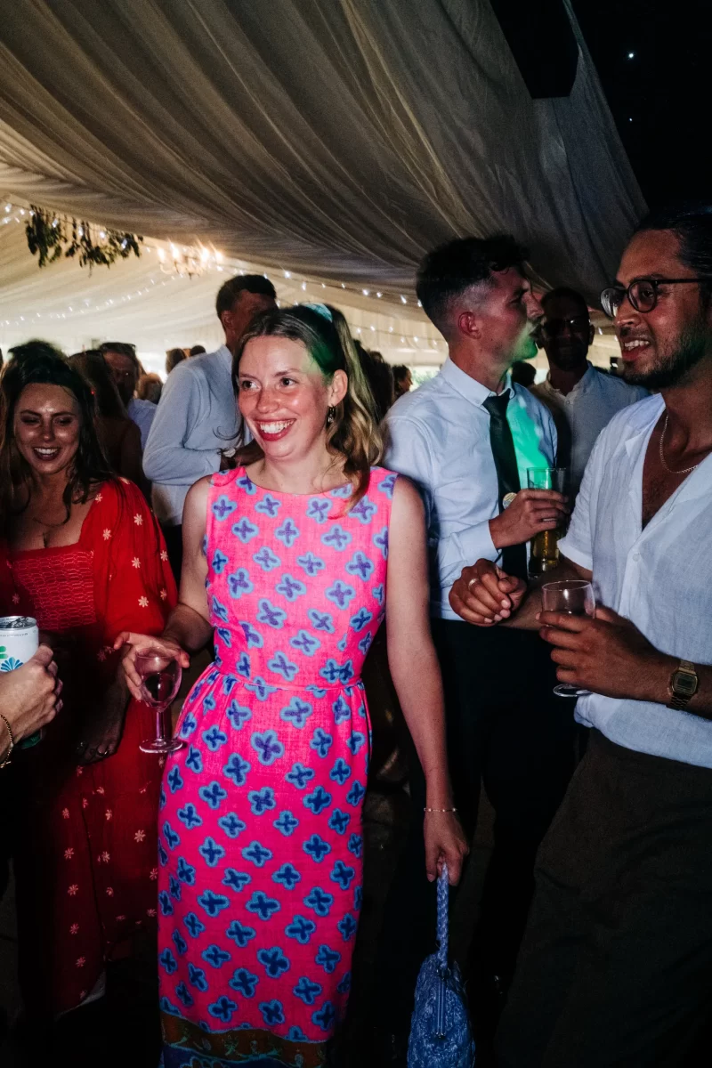 A woman in a bright pink and blue patterned dress smiles while holding a glass of wine at a lively indoor party, surrounded by people in festive attire—perfect inspiration for relaxed festival wedding or garden festival wedding ideas.