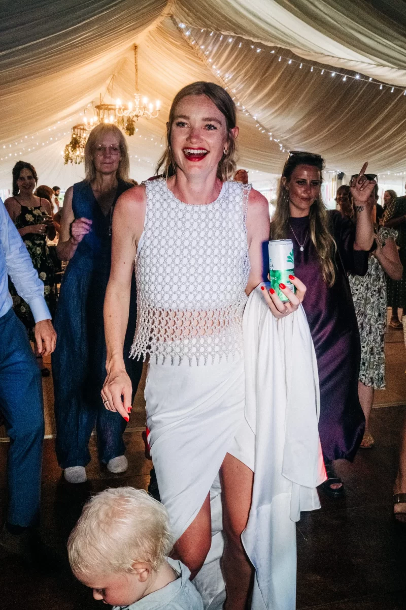 A smiling woman in a white top and skirt holding a drink walks through a lively crowd at a festively decorated indoor event, with string lights overhead and a small child in the foreground, capturing relaxed festival wedding vibes.