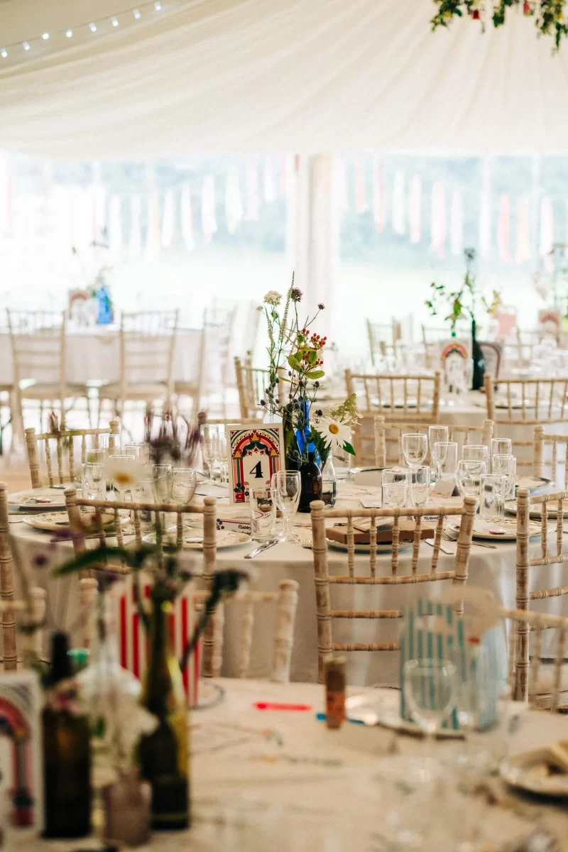 A bright, elegant wedding reception hall with round tables set with glassware, floral centerpieces, and table numbers. White chairs surround the tables, perfect for relaxed festival wedding vibes as light streams through a large, draped tent.
