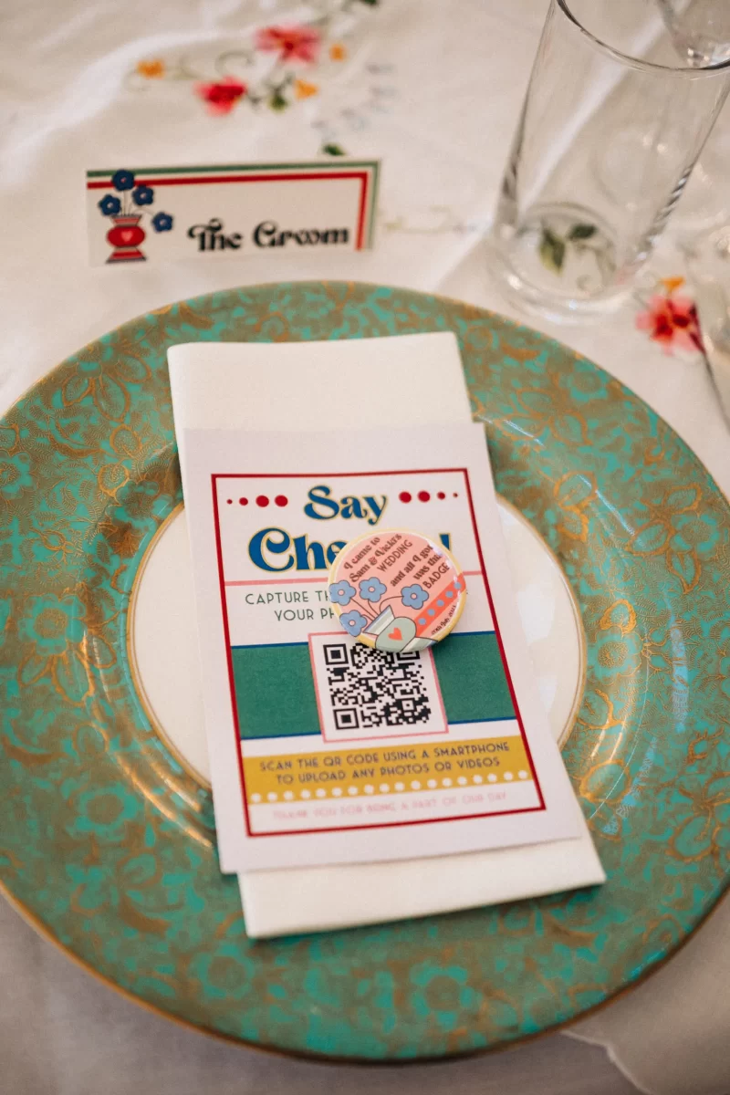 A decorative plate with a gold and teal pattern holds a folded napkin topped with a card and button. Perfect for relaxed festival wedding vibes, the card invites guests to upload photos via QR code. A name card labeled The Groom sits nearby.